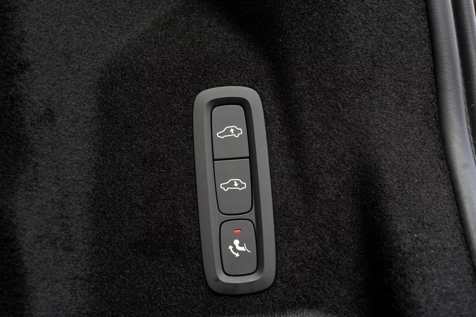 Detailný záber na ovládací panel vo vozidle Volvo XC60 2021 (2. generácia, facelift), zasadený v plastovom obložení proti čiernemu, koberčekovému pozadiu. Má štyri tlačidlá: hore - vozidlo s otvoreným kufrom, druhé - silueta auta so všetkými otvorenými dverami, tretie - auto so šípkou smerujúcou na kapotu, dole - auto s klenutou čiarou hore, pravdepodobne naznačujúcou uvoľnenie kufra, odomykanie/zamykaniu dverí, prístup ku kapote/režimu pre valetov a aktiváciu bezpečnostného systému alebo funkciu panic alarm.