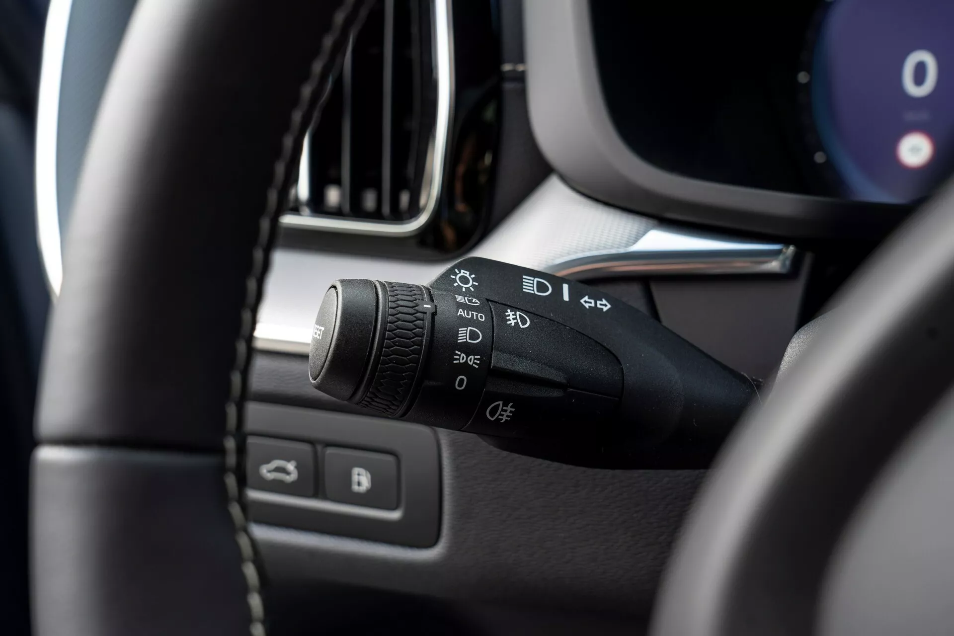 Detailný záber interiéru Volvo XC60 2021 (druhá generácia, facelift) so zameraním na ľavý stĺpik riadenia. Hlavným predmetom je ovládacia páka svetiel a smeroviek so symbolmi pre automatické svetlá, parkovacie svetlá, svetlomety, nastavenia hmloviek a prispôsobiteľnú funkciu. Obrázok je zhotovený z miesta vodiča, mierne nahor a doľava, ukazuje rozmazanú ľavú hornú časť volantu v popredí a zdôrazňuje ovládaciu páku. Ďalšie rozmazané detaily zahŕňajú časti prístrojového panelu, stredovej konzoly, okraju ventilácie alebo displeja informačného systému a dvere vodiča s ovládaním okien a zrkadiel.