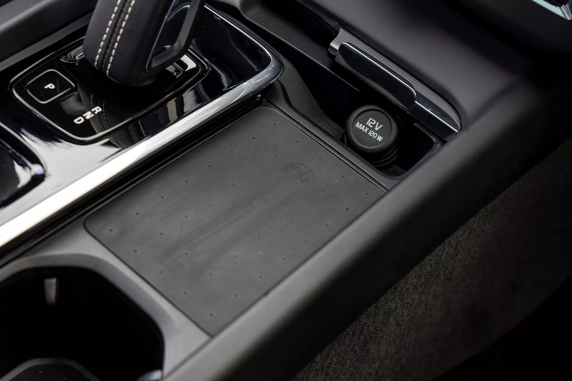 Detailný, šikmý pohľad na oblasť stredovej konzoly vo vozidle Volvo XC60 2021 (2. generácia facelift) z pohľadu spolujazdca, s automatickou radiacou pákou v polohe 'park', protišmykovým úložným priestorom, prepínačom elektronickej parkovacej brzdy, držiakmi na nápoje a kombináciou materiálov z mäkkého plastu a kovového povrchu v tlmených tmavošedých a čiernych tónoch.