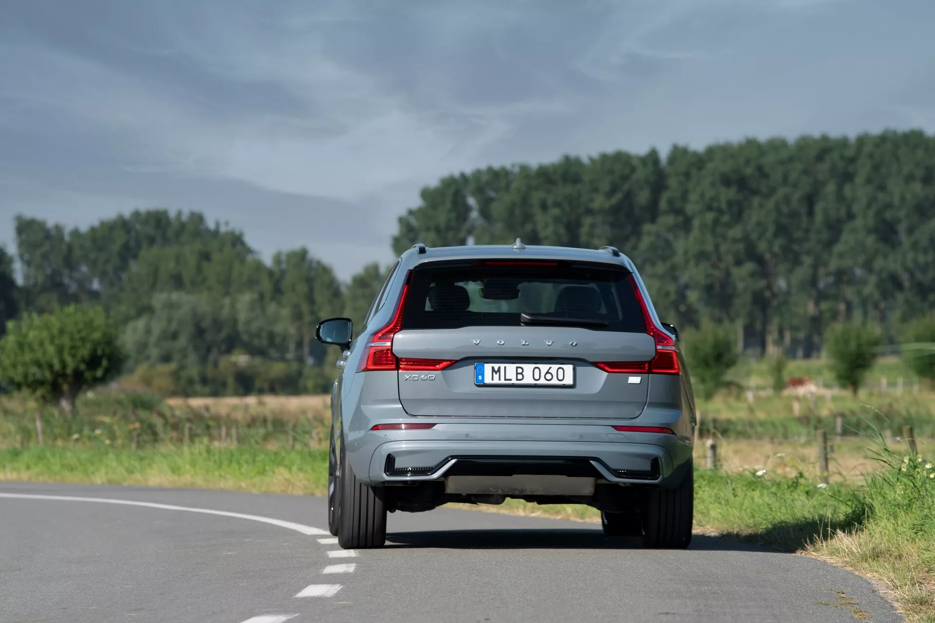 Zadný pohľad na moderné SUV 2021 Volvo XC60 (2. generácia-facelift) na dvojprúdovej ceste v prírodnom, vidieckom prostredí, zobrazujúce charakteristické zadné svetlá, dvojité výfuky, zadný spojler so zabudovanými brzdovými svetlami, cúvaciu kameru, zadný stierač a parkovacie senzory. Umiestnené mierne napravo od stredovej čiary, s čistou oblohou a stromami v pozadí.