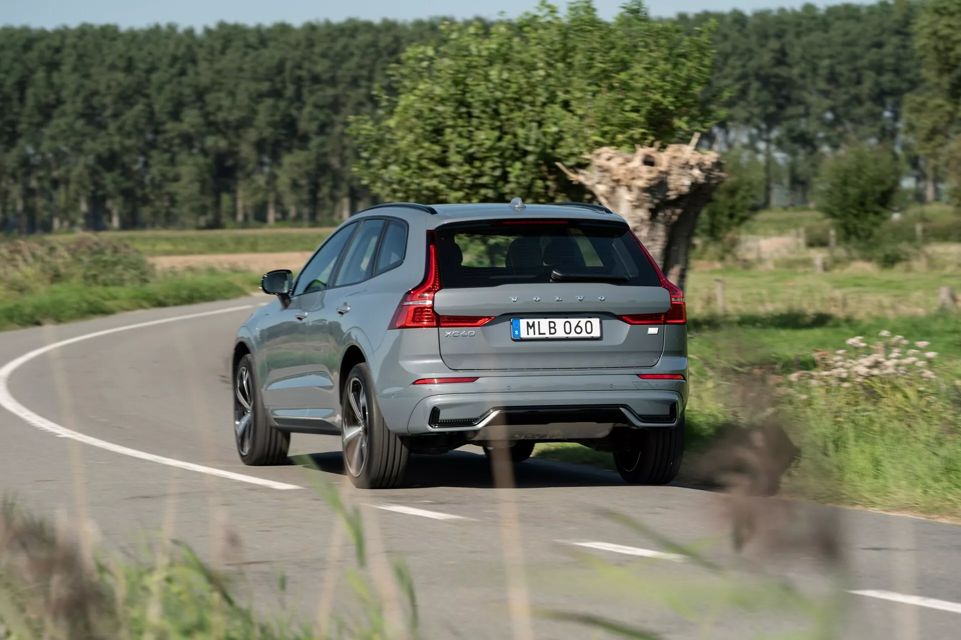 Zadný trojštvrťový pohľad na moderné kovovo šedé SUV Volvo XC60 2021 v pohybe na vidieckej ceste, zobrazujúci výrazné zadné svetlá, dvojitý výfukový systém, zadný stierač, výrazný zadný spojler, integrované senzory na zadnom nárazníku, centrálne umiestnenú ŠPZ, liatinové kolesá, ostrú líniu pozdĺž boku, reflexné okenné lišty a strešný systém. Pozadie s rozmazaným lístím a jasnou oblohou naznačuje pohyb a vidiecke prostredie.