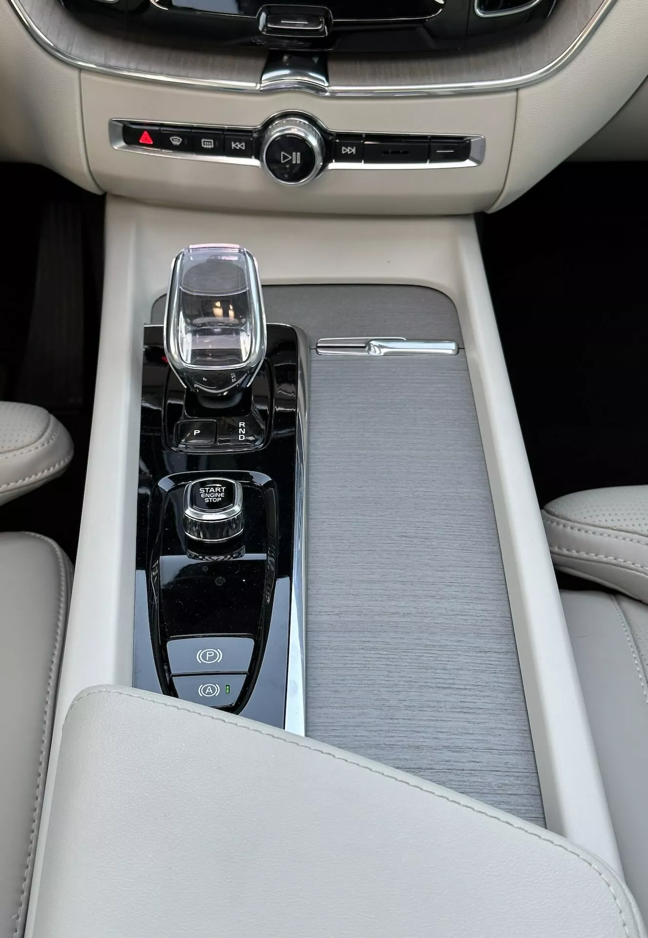 Detailný pohľad na stredovú konzolu vo Volvu XC60 Recharge plug-in hybrid z roku 2021, s béžovou koženou lakťovou opierkou, posuvným kartáčovaným povrchom kovu či plastu, moderným ovládačom infotainmentu, kryštálovým voličom prevodovky pre automatickú prevodovku a ovládacími prvkami pre start/stop motora, vypnutie trakčnej kontroly a režim jazdných dynamík, spoločne s ovládaním klimatizácie pre dve zóny.