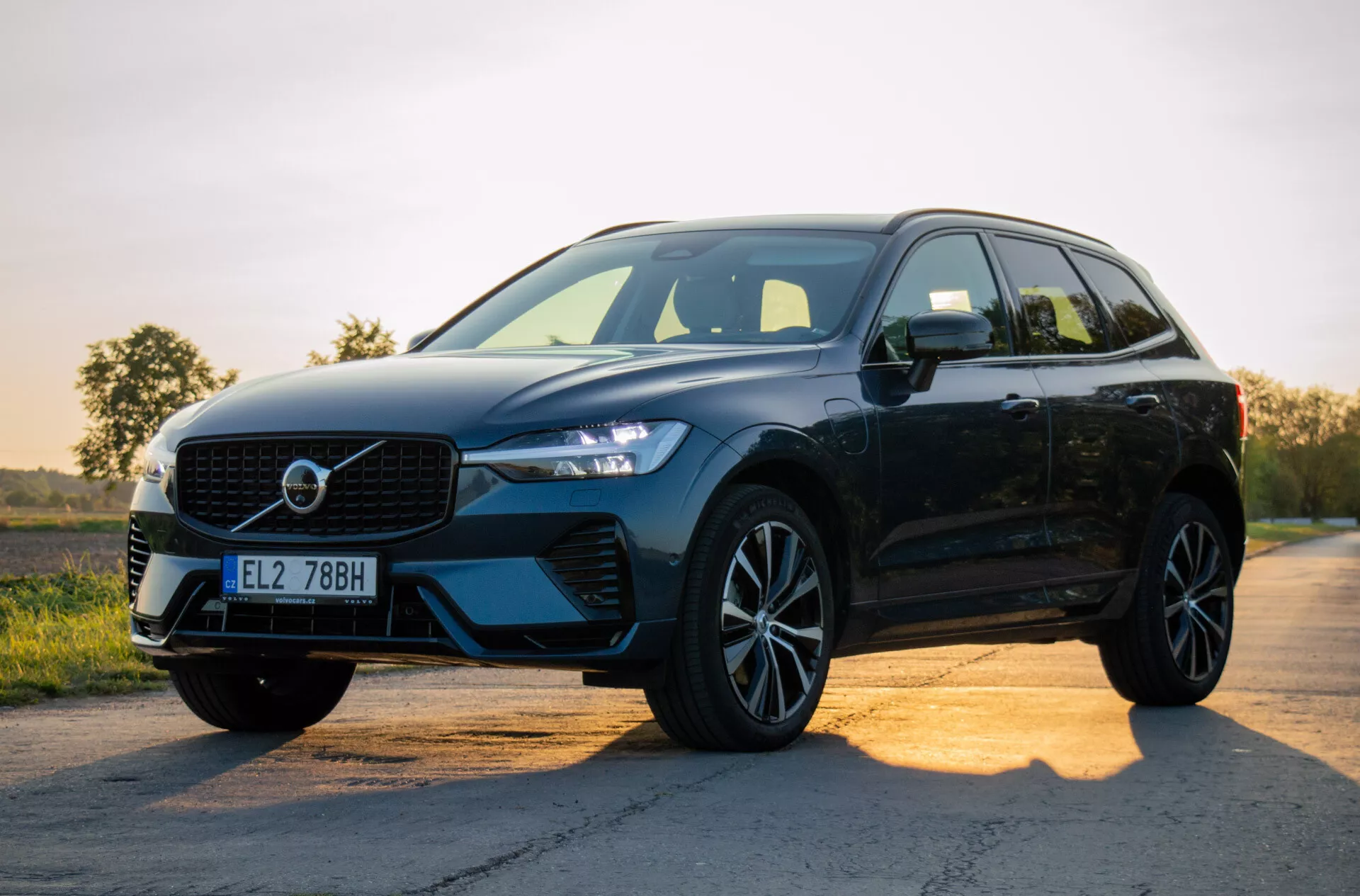 Čierne moderné SUV Volvo XC60 Recharge Plug-in Hybrid 2021 zaparkované na asfaltovej ceste pri východe alebo západe slnka, predstavujúce jeho dizajn s dôrazom na mriežku, LED svetlomety, zliatinové kolesá a bočný profil na pozadí gradientového neba