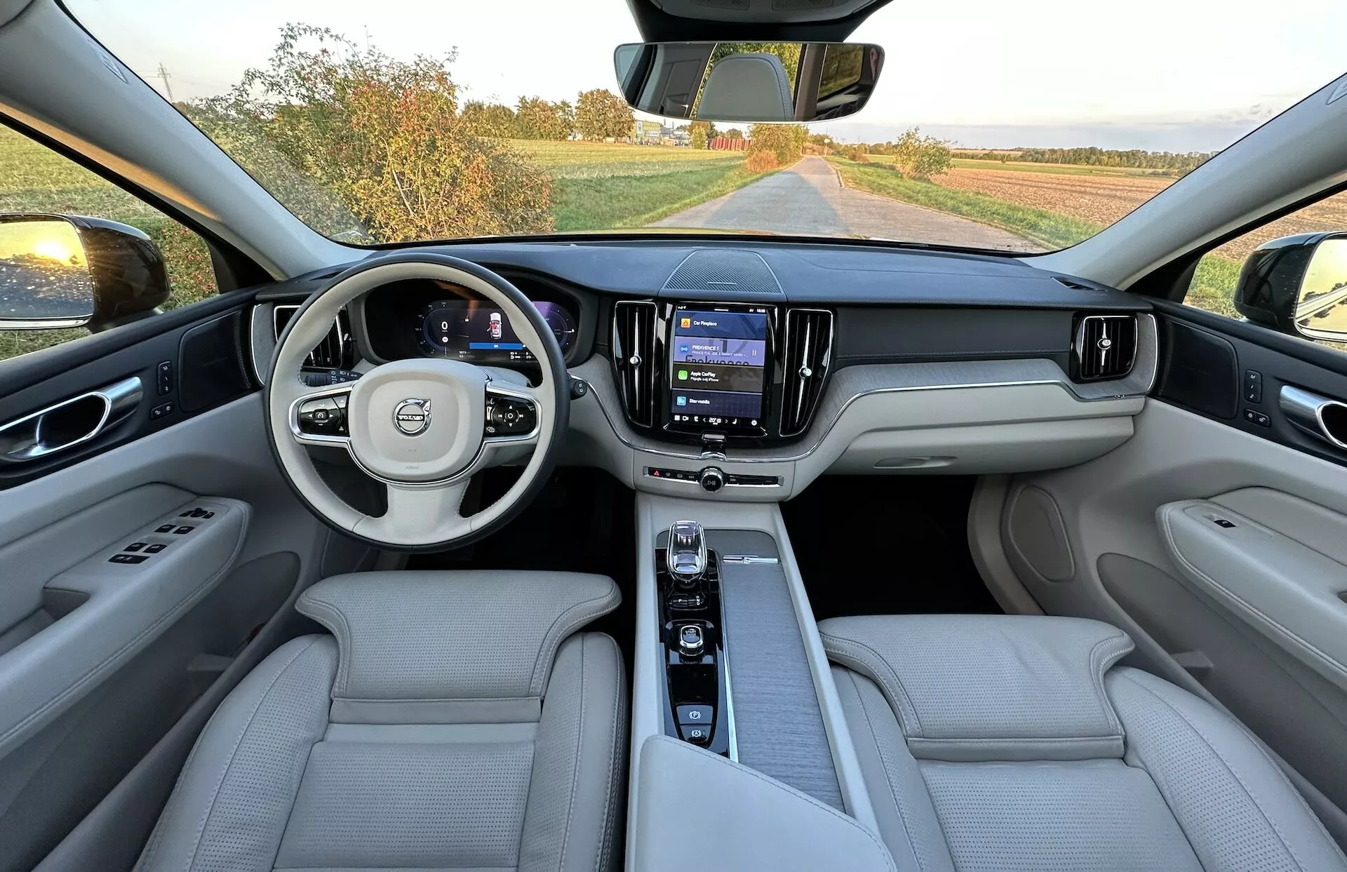 Interiérový pohľad na Volvo XC60 Recharge Plug-In Hybrid 2021 z oblasti zadných sedadiel zobrazujúci celú prístrojovú dosku, multifunkčný volant s logom, digitálny panel prístrojov, centrálny dotykový infotainment systém a luxusné sedadlá z bledokoženého čalúnenia.