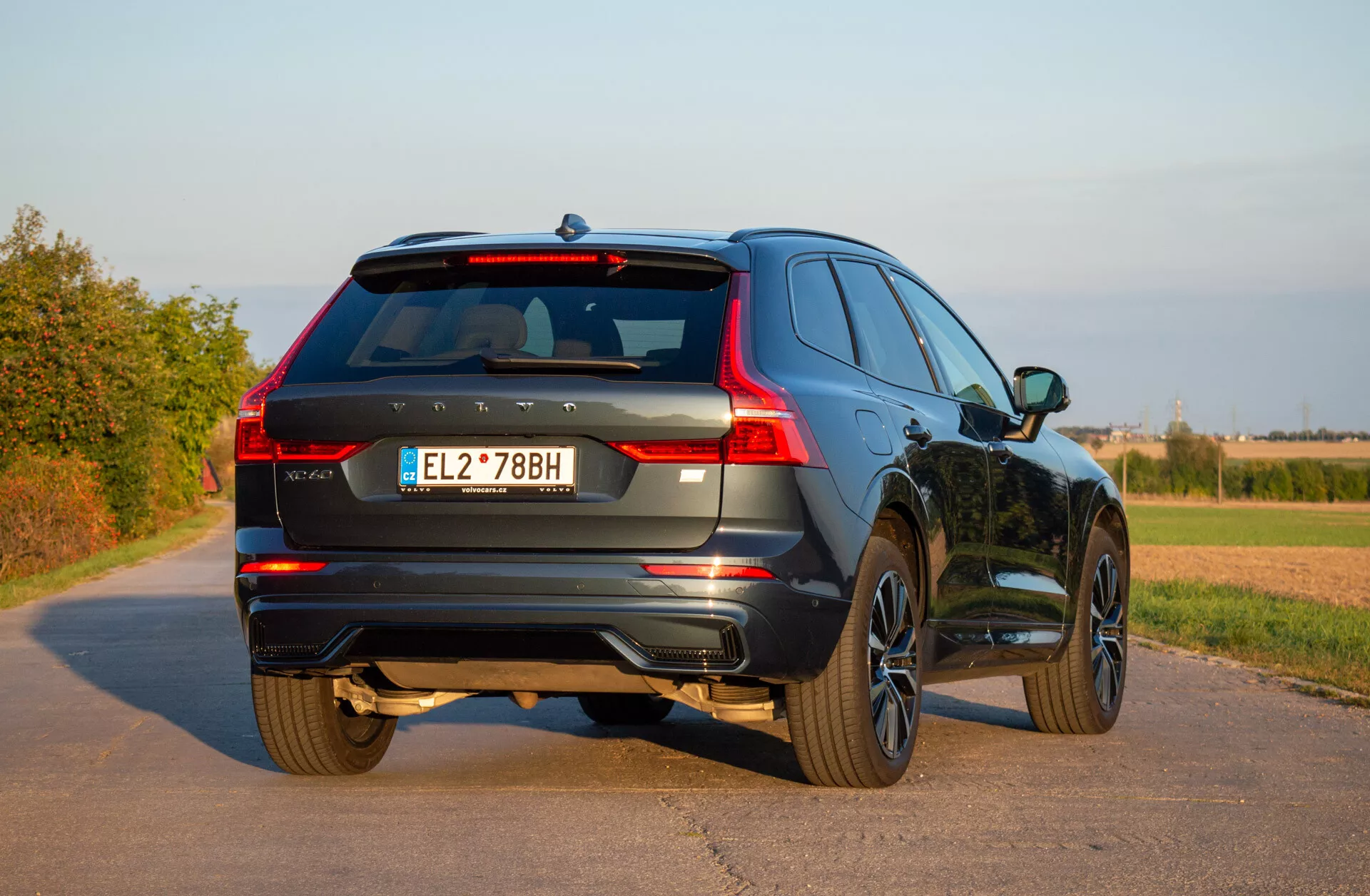 Stacionárne SUV Volvo XC60 Recharge plug-in hybrid z roku 2021 na spevnenej ceste ukazujúce svoj zadný trojštvrťový pohľad, zdôrazňujúce LED zadné svetlá, zliatinové kolesá a dynamický dizajn v rurálnom prostredí pod jasnou oblohou