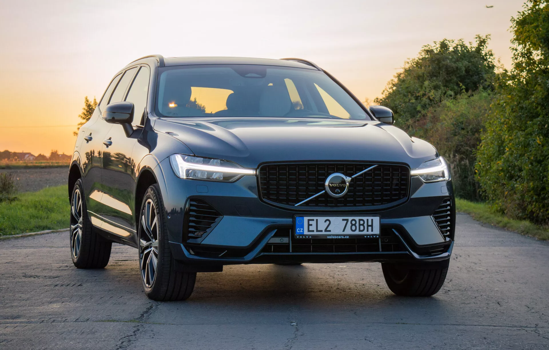 Luxusné SUV Volvo XC60 Recharge Plug-In Hybrid 2021 na zpevnenej ceste s prírodnou krajinou počas zlatej hodinky, ukazujúce prednú mriežku, LED svetlomety, tvarovanú kapotu, viacramenné zliatinové kolesá a lesklý povrch