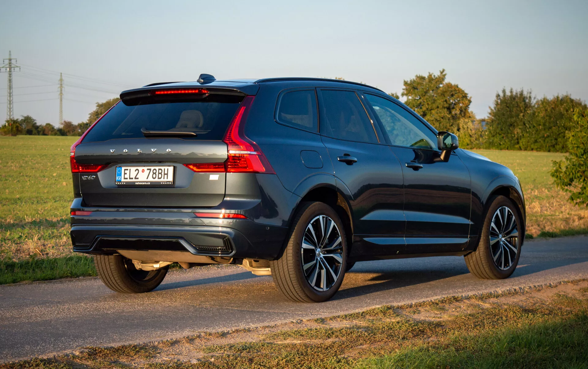 Volvo XC60 Recharge Plug-In Hybrid 2021 v hlbokomodrej, predvádzajúce aerodynamický dizajn a moderné prvky ako sú LED zadné svetlá, zliatinové kolesá a dvojitý výfukový systém, zaparkované na vidieckej ceste pod jasným nebom
