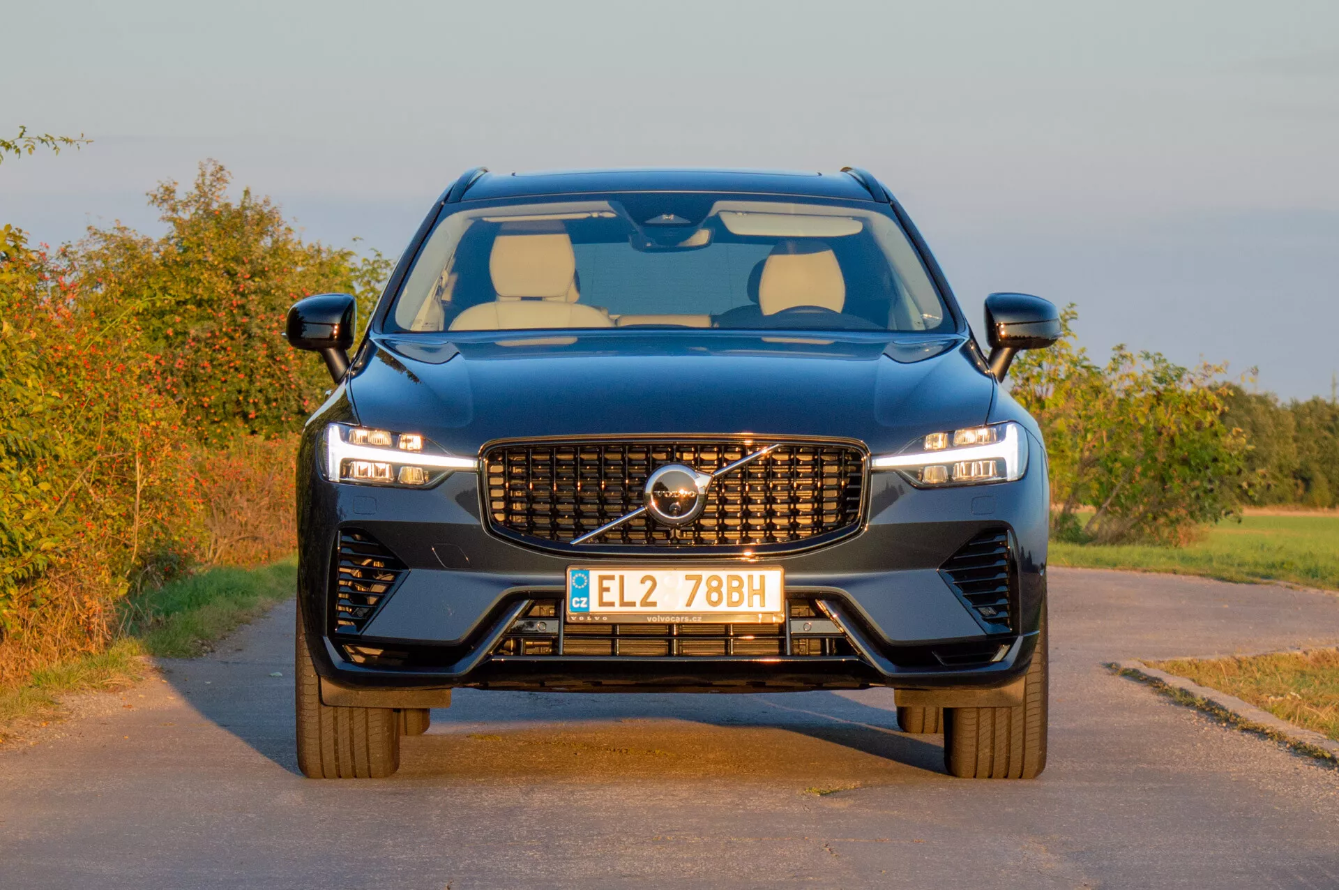 Volvo XC60 Recharge z roku 2021 predvádza svoj moderný dizajn s charakteristickou mriežkou, štíhlymi LED svetlometmi a športovým predným nárazníkom na pozadí vidieckej krajiny v teplom svetle neskorého popoludnia