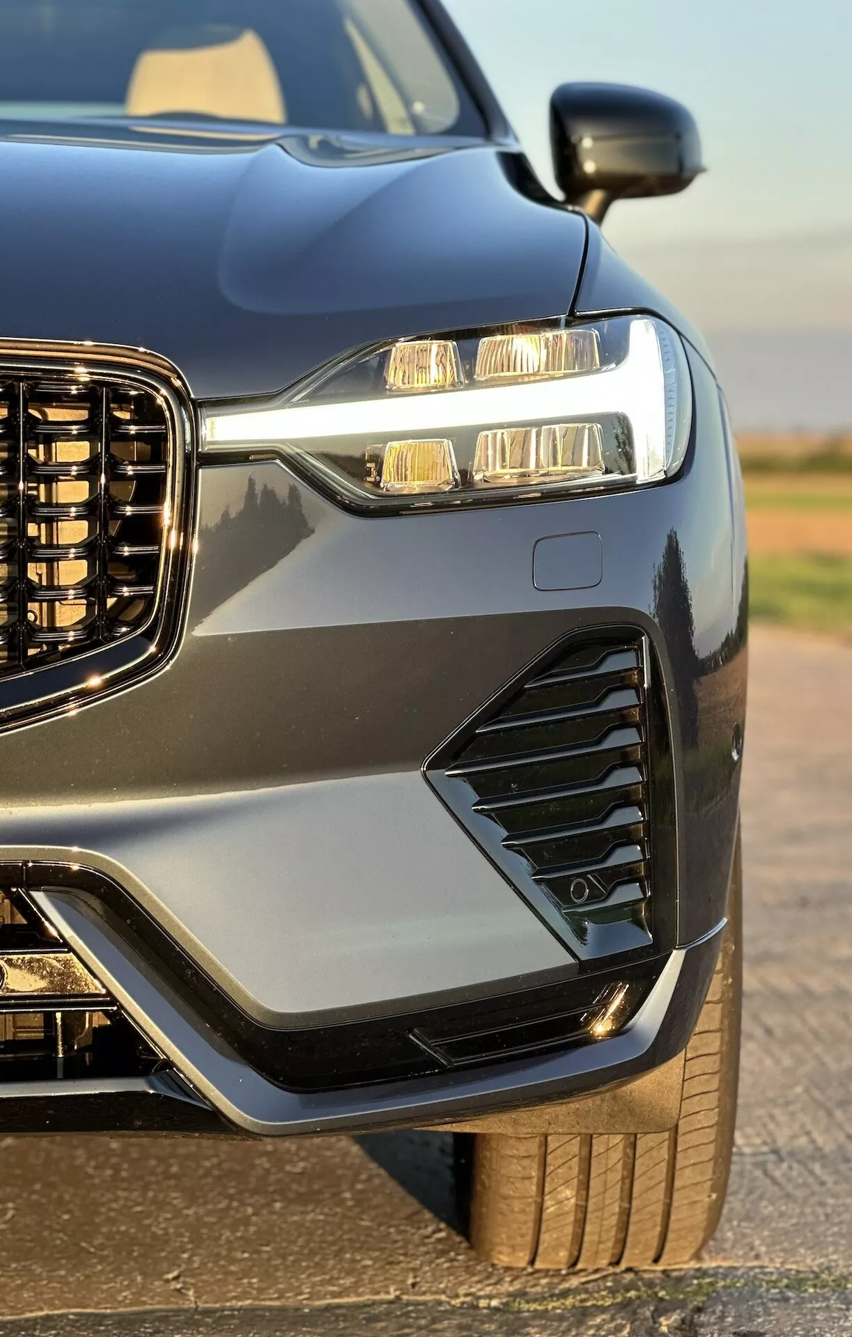 Detailný pohľad z prednej časti a boku na Volvo XC60 Recharge Plug-In Hybrid z roku 2021, zdôrazňujúci jeho vertikálnu mriežku, LED svetlomety s dennými svetlami, štýlové ventilácie a hmlové svetlá, športovú kapotu a blatníky, metalický lak s kontrastným lemovaním a náhľad na jeho robustné koleso a pneumatiku, zvýraznený lesklým povrchom vozidla.