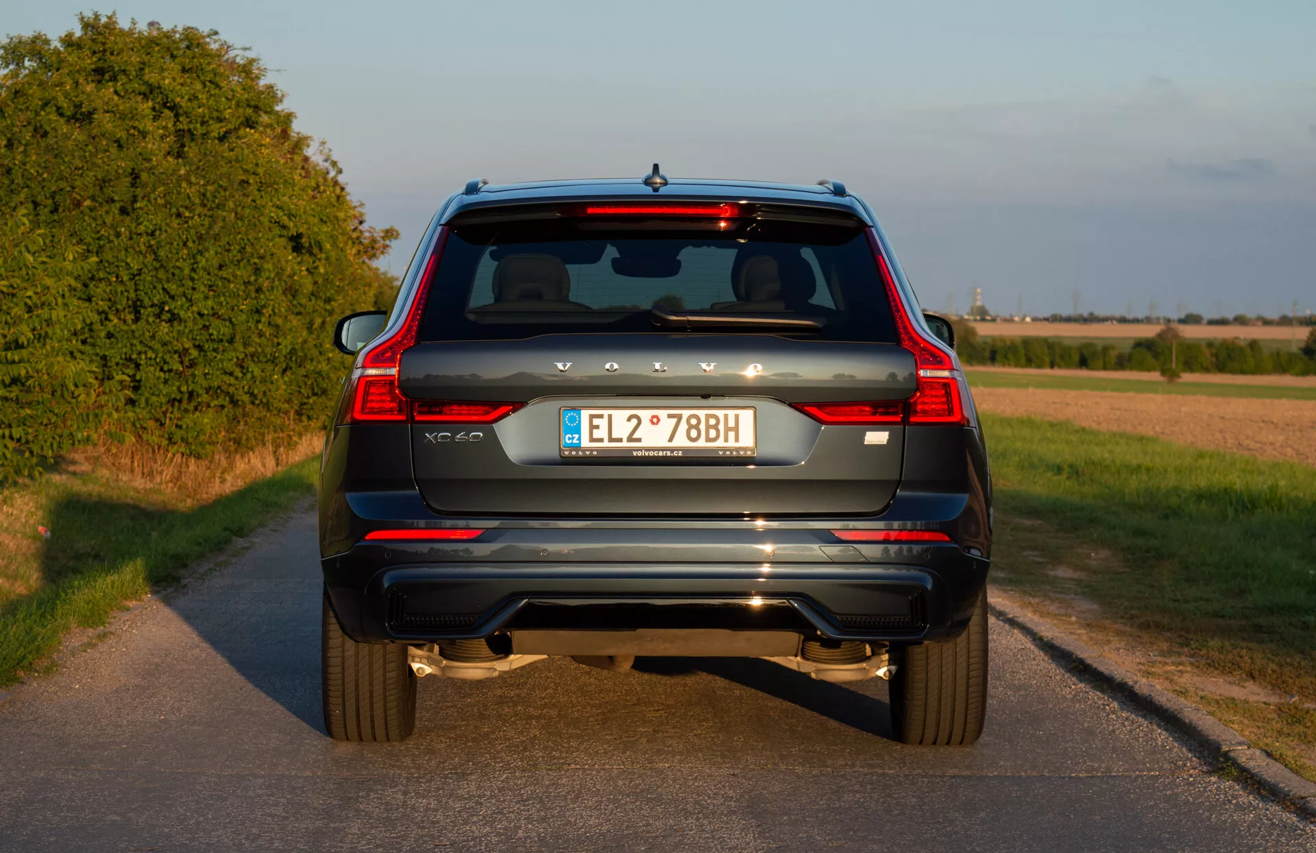 Zadný pohľad na modré SUV Volvo XC60 Recharge Plug-In Hybrid z roku 2021 so zvislými zadnými svetlami, dvojitými výfukovými koncami, zadnými odrazkami, stieračom zadného okna, tretím brzdovým svetlom, európskou poznávacou značkou a zadnou kamerou na vidieckej ceste pred západom slnka