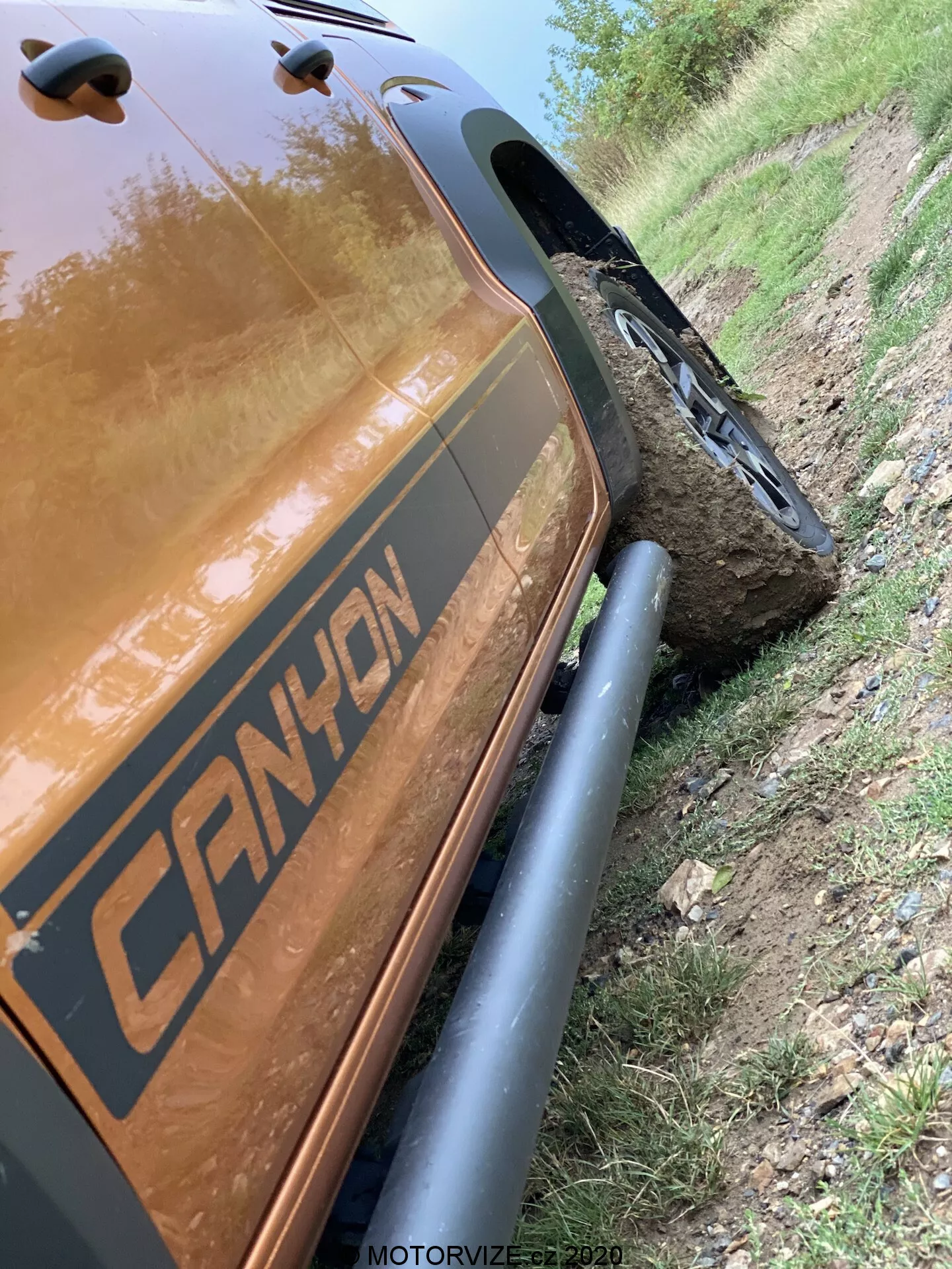 Tento naklonený obraz pod uhlom 45 stupňov zobrazuje zadnú časť na strane spolujazdca VW-Nutzfahrzeuge Amarok 2016. Zameriava sa na detailný záber od zadného blatníka po zadnú nákladnú časť, zdôrazňujúc robustné čierne ochranné kryty blatníkov a robustné terénne pneumatiky. Vozidlo má grafiku s nápisom 'CANYON' a je zaparkované vedľa prašnej cesty s kolesom namontovaným na veľkom kameni, čo demonštruje jeho terénne schopnosti. Reflexná metalická hnedá alebo medená farba karosérie je výrazná, s odleskmi svetla. Časová značka 'MOTORVIZE.cz 2020' naznačuje, že bola zachytená pre motoristickú publikáciu.