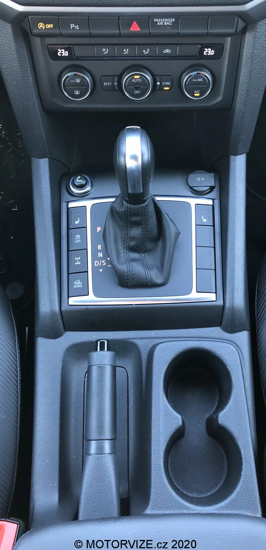 Tento obrázok ukazuje stredovú konzolu vw-nutzfahrzeuge amarok 2016 z nadhľadového uhla, približne z pohľadu vodiča alebo predného spolujazdca. Fotografia zachytáva interiér auta špecifický pre radiacu páku, ovládanie stredovej konzoly a úložné priestory. Obrázok zahŕňa ovládacie prvky klimatizačného systému, koženú riadiacu páku zasadenú do strieborného panelu naznačujúceho automatickú prevodovku, riadenie elektronickej parkovacej brzdy, úložný priestor, dva držiaky nápojov a ďalší úložný priestor smerom k zadným sedadlám. Viditeľné materiály zahŕňajú plast, kovové akcenty a kožu, ktoré odrážajú kombináciu funkčnosti a estetiky.