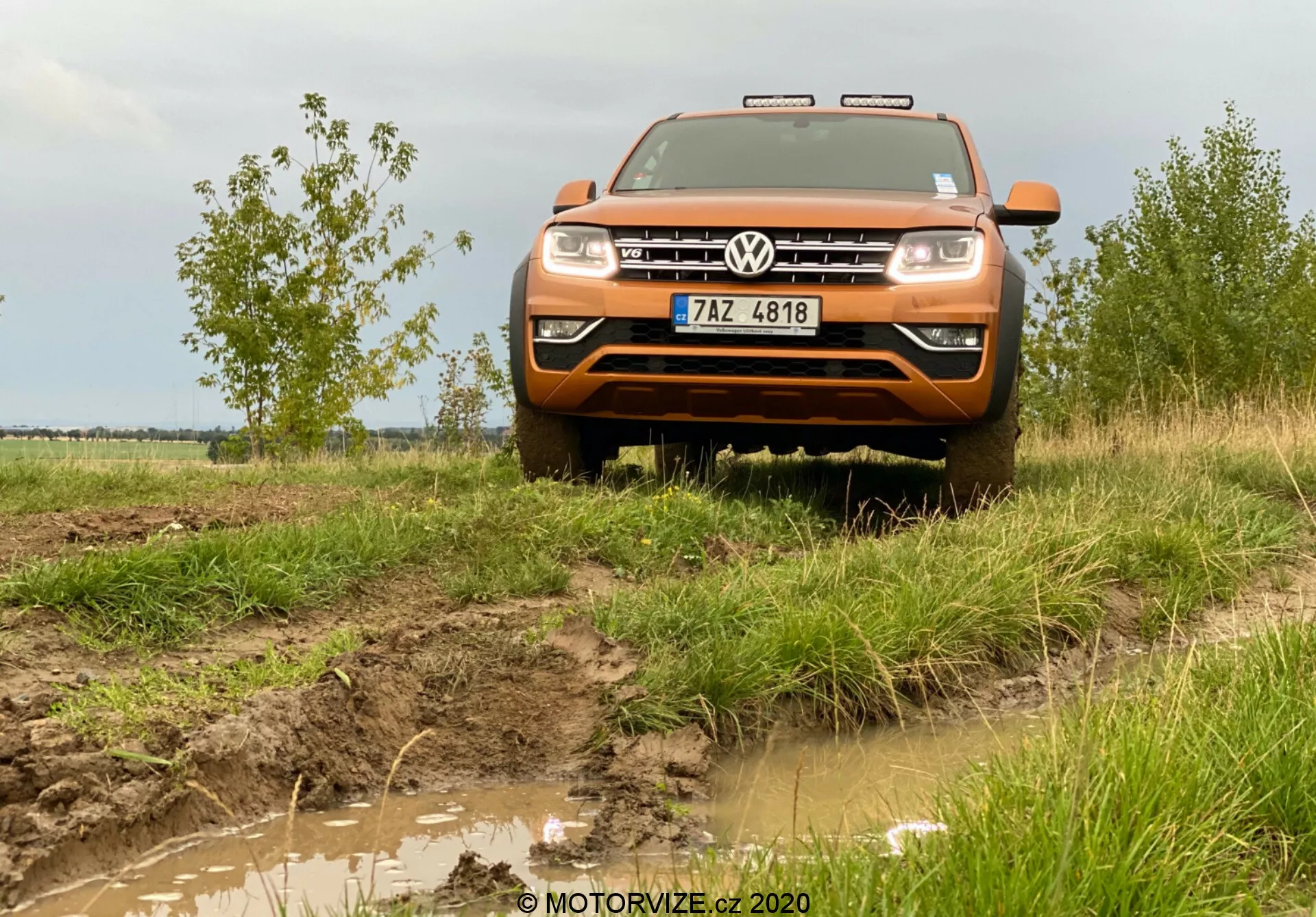 Čelný pohľad z mierne nízkeho uhla na oranžový Volkswagen Amarok 2016, stredne veľké pickup vozidlo, známe pre svoje terénne schopnosti. Obrázok zachytáva predok vozidla vrátane kapoty, mriežky, nárazníka, časti blatníkov, svetlometov a prominentne umiestneného loga Volkswagen v strede mriežky. Nad mriežkou sú vidieť chrómové akcenty alebo LED denné svietenie. Blatistý terén a tráva okolo vozidla zdôrazňujú jeho terénny charakter, s dodatočnými svetlami na streche, predným ochranným krytom a vidieckym pozadím s oblačnou oblohou dotvárajú adventúrne prostredie. Obrázok je zhotovený z približne dvoj- až trojmetrovej vzdialenosti, čím poskytuje detailný pohľad na predné časti vozidla a jeho interakciu s prostredím.