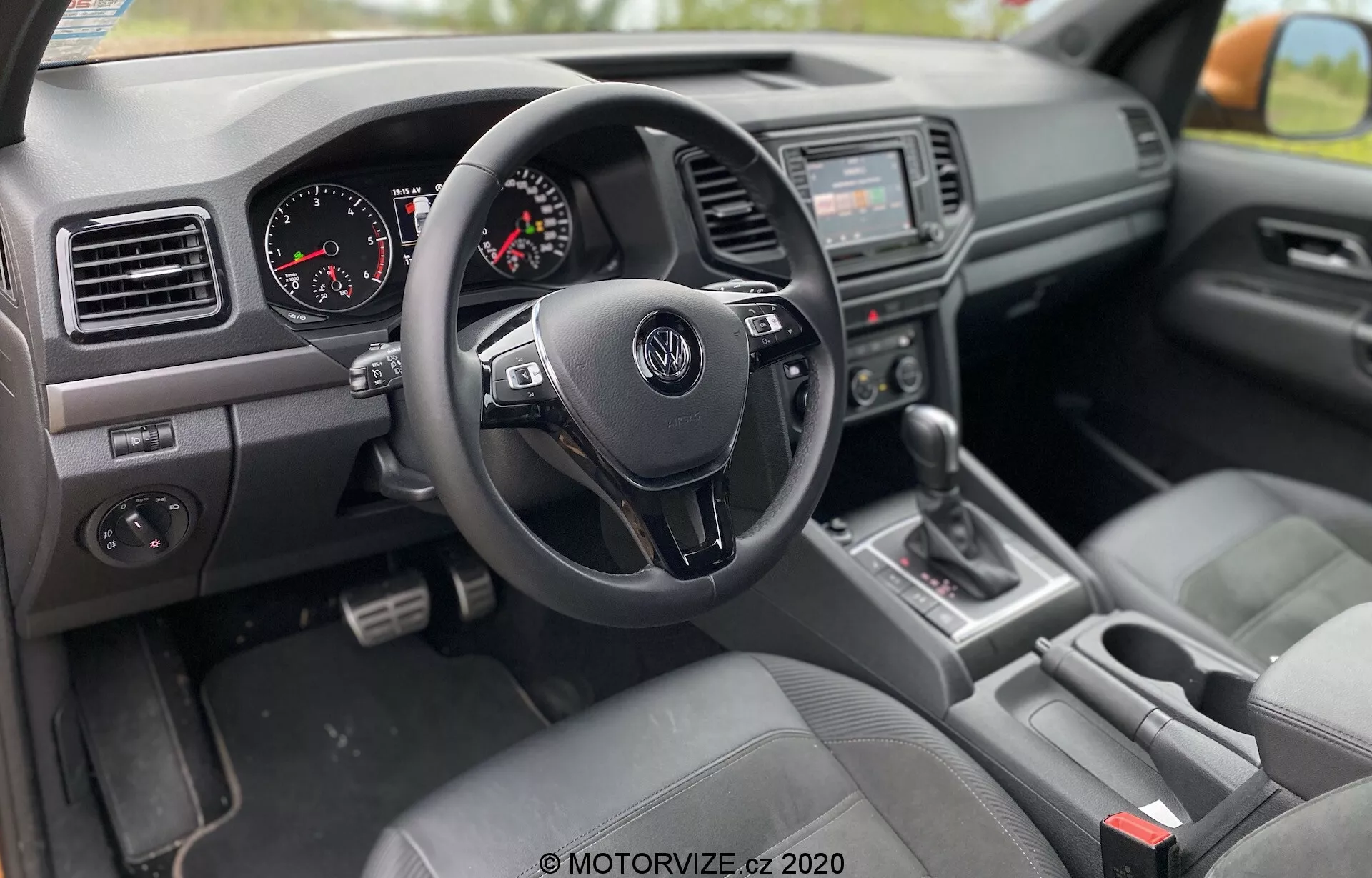 Pohľad na vnútro modelu VW Nutzfahrzeuge Amarok 2016 zo strany spolujazdca, s pravostranným riadením, trojramenným volantom Volkswagen s ovládacími tlačidlami, prístrojovou doskou s analógovým rýchlomerom a otáčkomerom, stredovou konzolou so zvukovým systémom a infotainment obrazovkou, radiacou pákou v polohe 'P', sedadlami s látkovým poťahom, prístrojovou doskou s ventilačnými otvormi a textúrovanými plastmi a ovládacím panelom okien a zrkadiel na dverách vodiča.