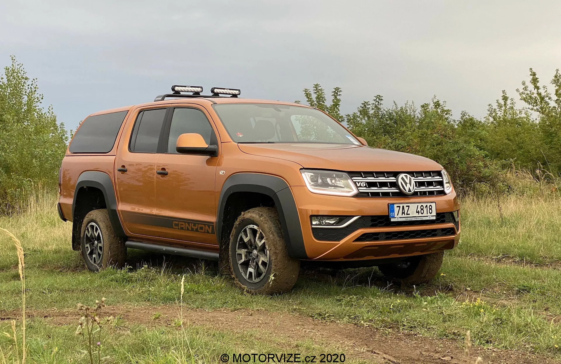 Obrázok zobrazuje oranžový pickup Volkswagen Amarok 2016 zaparkovaný na nerovnom trávnatom teréne s pozadím oblačnej oblohy. Pohľad zo strany prednej časti sa zameriava na stranu vodiča, ukazuje profil vozidla a čiastočný pohľad na prednú časť. Kľúčové viditeľné prvky zahŕňajú strešný nosič, kolesá pokryté blatom naznačujúce nedávnu terénnu aktivitu, bočné stupne, konfiguráciu s dvojitou kabínou, moderný dizajn svetiel a predný nárazník s hmlovými svetlami a spodným ochranným krytom. Vozidlo vyžaruje drsnú terénnu úžitkovú estetiku s modernými technologickými prvkami.