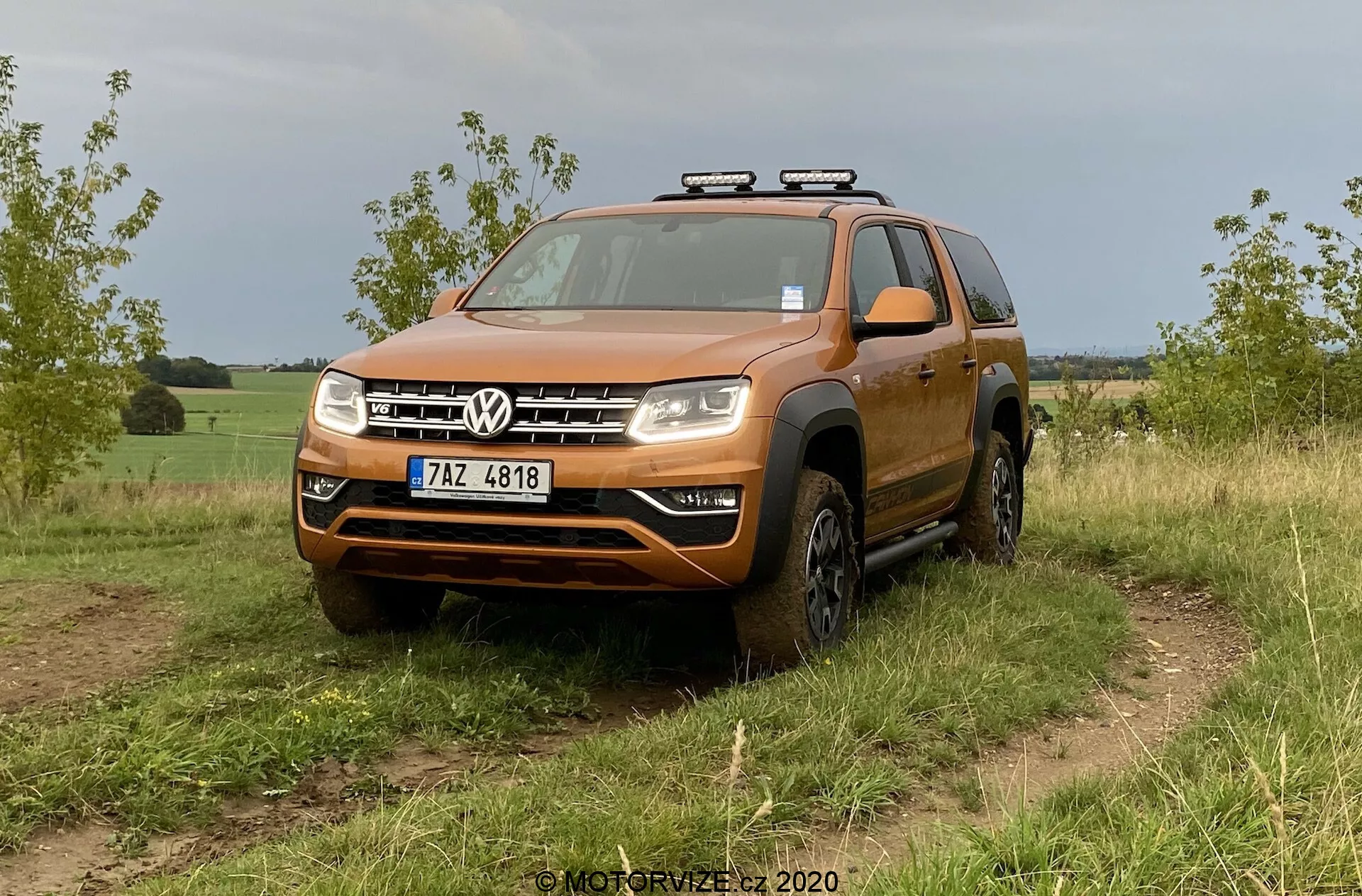 Oranžové SUV Volkswagen, konkrétne model VW Amarok 2016, je zobrazené na trávnatom teréne. Snímka z nízkeho až stredného pohľadu zpredu zdôrazňuje robustný dizajn vhodný na off-road aktivity. Vozidlo má výraznú prednú mriežku s dvoma chrómovanými pruhmi ohraničujúcimi centrálne logo Volkswagen, značný nárazník s možnou ochrannou doskou, číre kompozitné svetlomety s potenciálnymi LED alebo halogénovými žiarovkami a rozšírené oblúky kolies s off-road pneumatikami. Na streche sú namontované pomocné svetlá, čo naznačuje väčšiu funkčnosť pre nočnú jazdu. Viditeľné detaily zahŕňajú čelné sklo, strešné lišty, spätné zrkadlá s integrovanými smerovkami a prednú registračnú značku. Vozidlo vyzerá čisto s trochu špiny na dolných častiach a kolesách, čo zodpovedá off-road použitiu. Pozadie naznačuje vidiecke alebo polovidiecke prostredie.