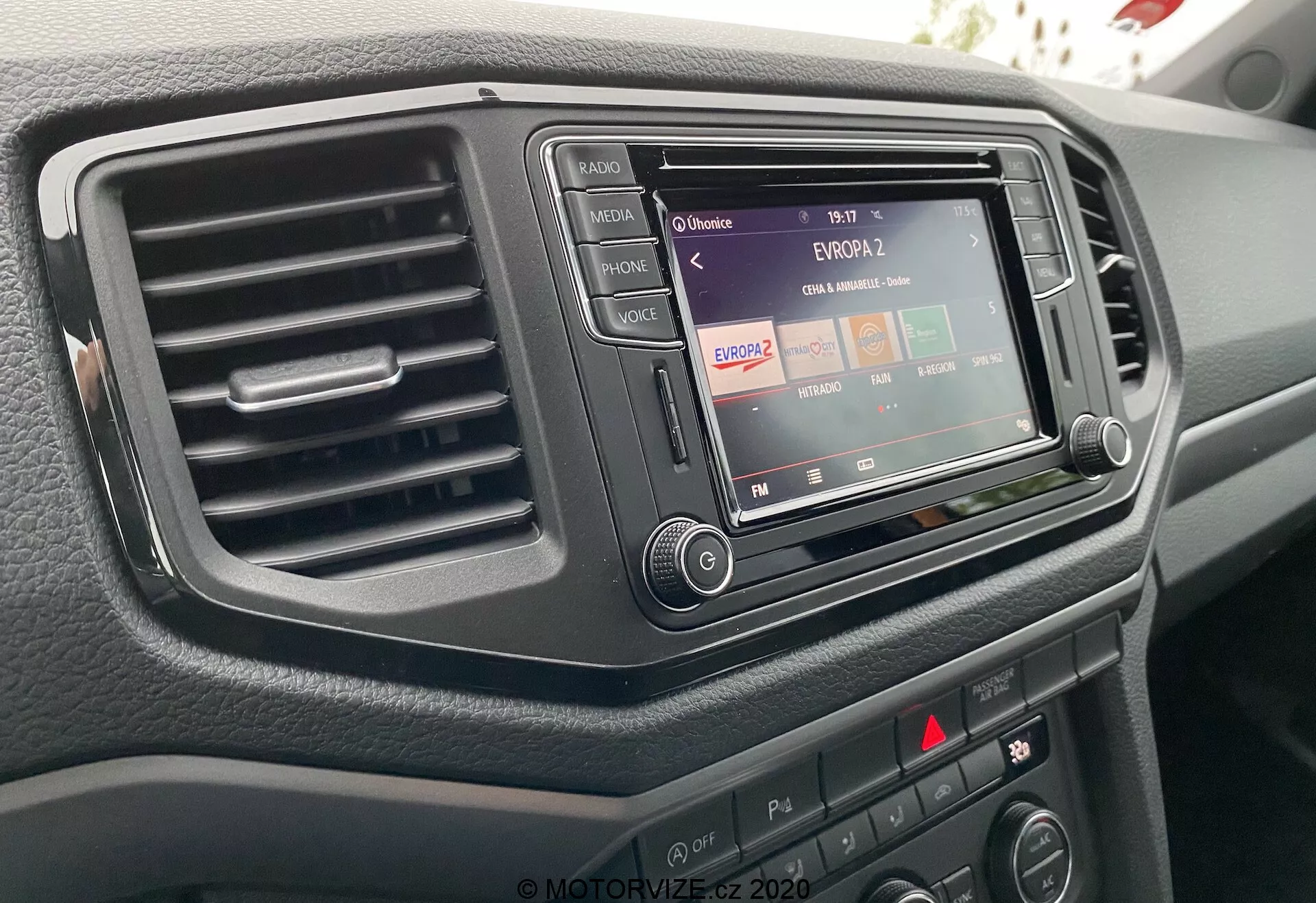 Detailný záber na stredovú konzolu VW Nutzfahrzeuge Amarok 2016 z pohľadu vodiča, zvýrazňujúci infotainment systém s rádiovou funkciou, jedným výduchom klimatizácie vľavo a rôznymi ovládacími tlačidlami a gombíkmi. Infotainment systém je zasadený do tmavého, elegantného rámu a povrch palubnej dosky má textúrovanú úpravu.