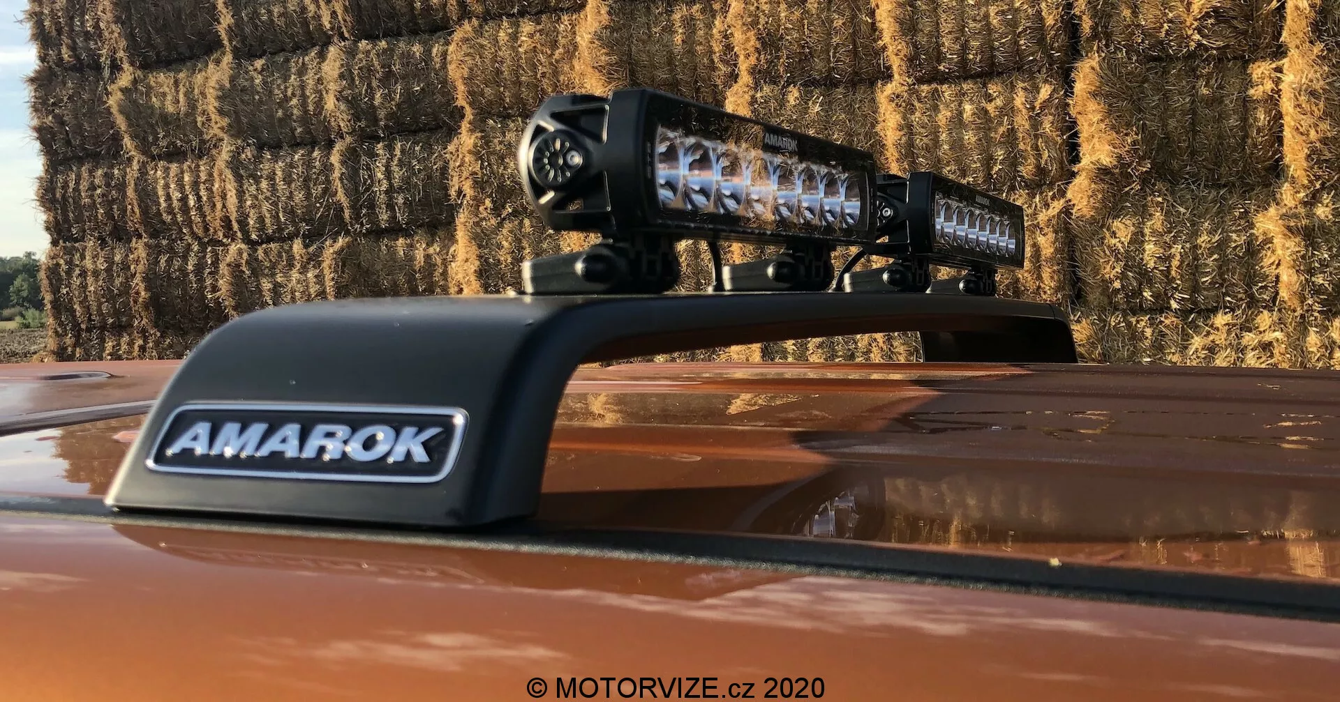 Detailný záber na pickup Volkswagen Amarok 2016 s namontovaným aftermarket LED svetelným panelom na streche. Čierne púzdro svetelného panelu s viditeľnými jednotlivými LED prvkami nesie označenie 'AMAROK' na vetracej krytke. Fotografia bola vytvorená vonku a ukazuje reflexné šošovky svetelného panelu a balíky slamy v pozadí, čo naznačuje vidiecke prostredie.