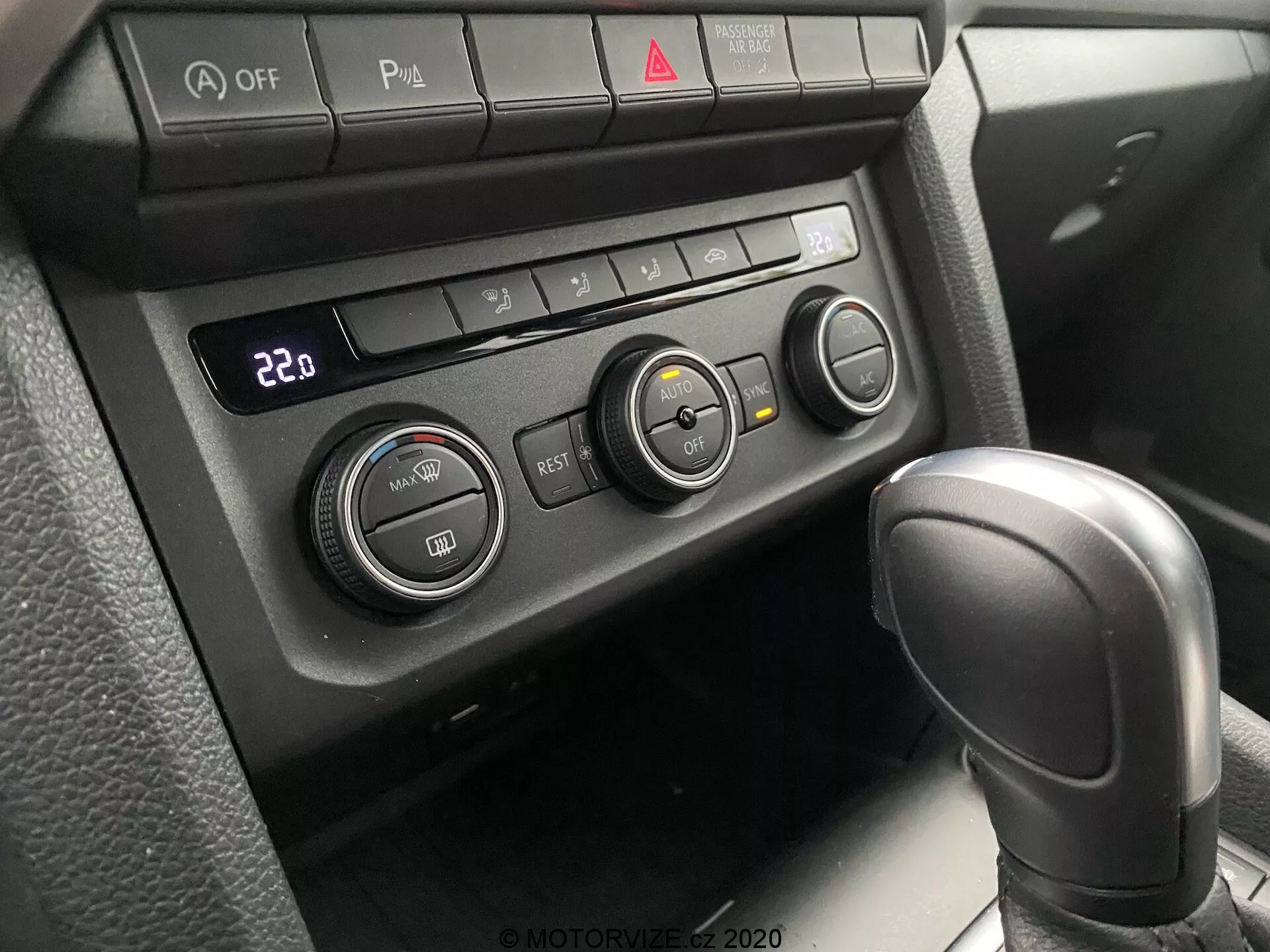 Zblízka pohľad na klimatizačný systém a čiastočnú radiacu páku VW Nutzfahrzeuge Amarok 2016. Perspektíva pasažiera alebo vodiča sústredeného smerom nadol na stredovú konzolu. Obsahuje koženú automatickú radiacu páku v polohe 'Park' a klimatizačný panel nastavený na 22°C s tromi otočnými ovládačmi a niekoľkými funkčnými tlačidlami. Tlačidlá zahŕňajú vypnutie Auto Stop/Start, prepínač parkovacích senzorov, vyhrievaný volant a vyhrievané sedadlo spolu s červeným tlačidlom výstražných svetiel uprostred. Mäkké a rozptýlené osvetlenie naznačuje dobre zatienený interiér.