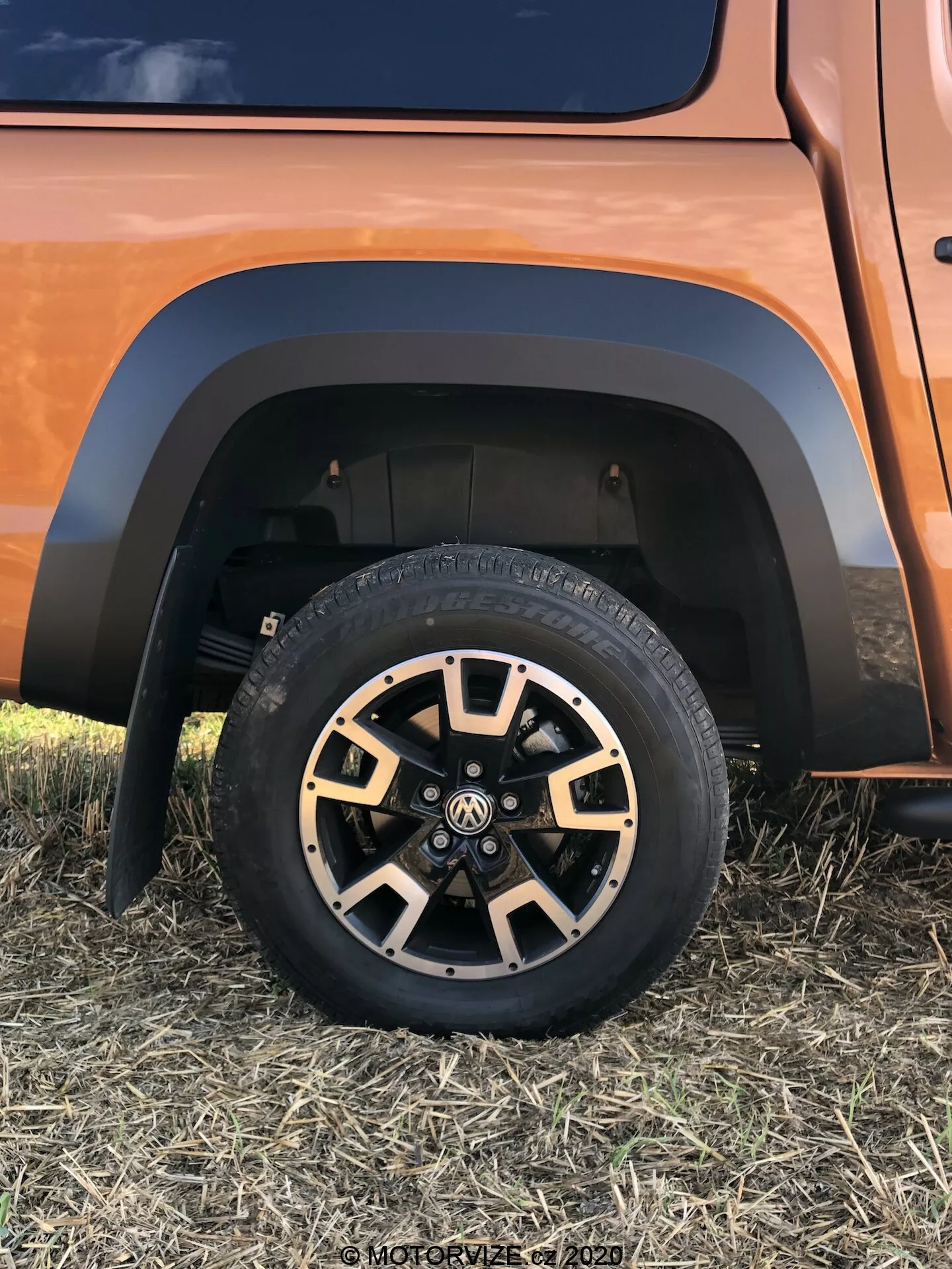 Detailný pohľad na spodnú časť vozidla VW Nutzfahrzeuge Amarok 2016, zobrazujúci robustnú terénnu pneumatiku na viaclúčovom zliatinovom kolese s čiernymi a kovovými povrchovými úpravami, kontrastný blatník a časť odpruženia v zemitej alebo medenej farbe vozidla, zo snímky urobené z nízkeho bočného uhla spredu.