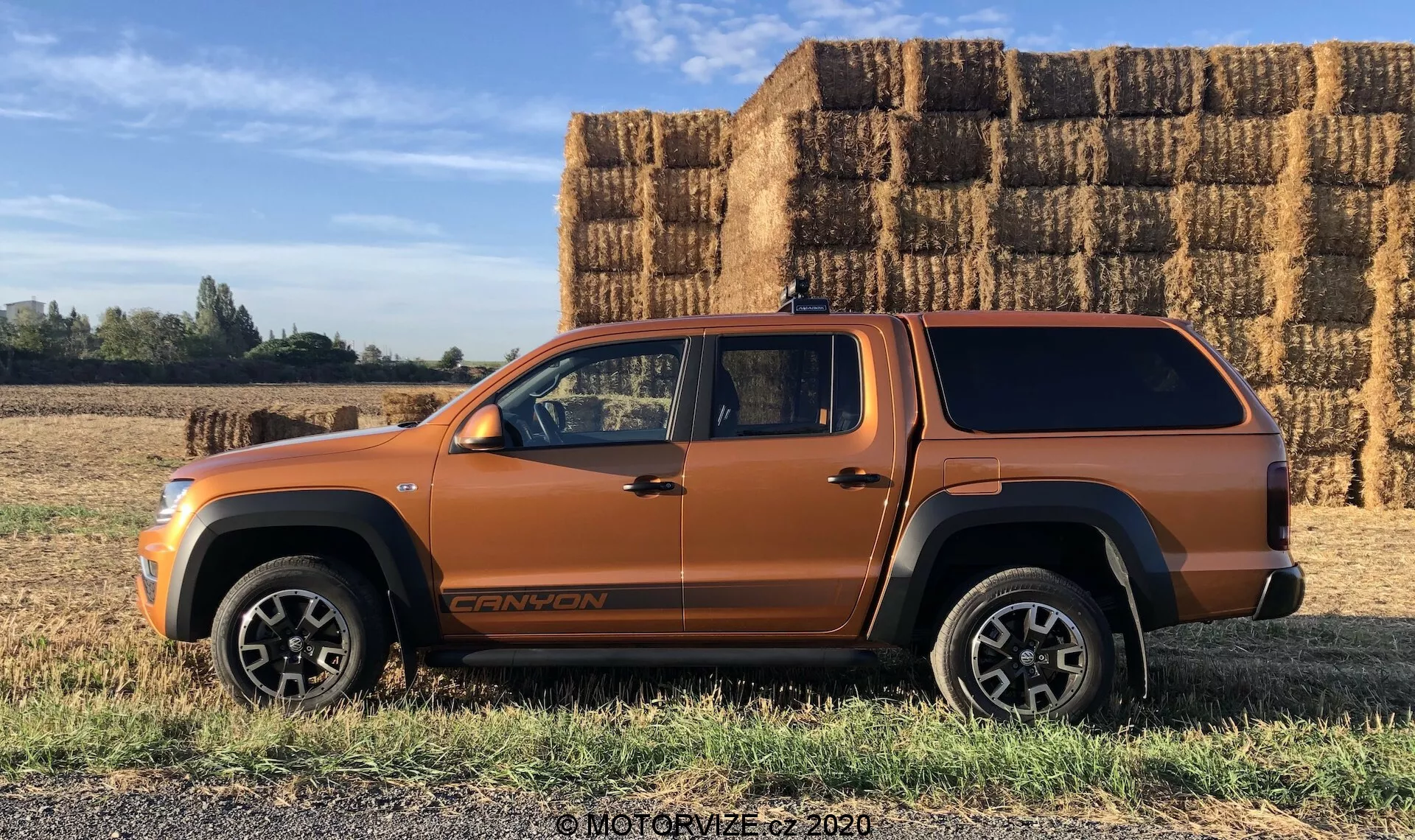 Bočný profil metalického oranžového SUV typu vozidla, pravdepodobne stredne veľkého SUV alebo pickup s predĺženou kabínou a krytom ložného priestoru, zaparkovaného na rovnej trávnatej ploche s veľkými obdĺžnikovými balíkmi sena v pozadí pod jasnou oblohou s jasným slnkom. Vozidlo má výrazné línie karosérie, veľké oblúky kolies, čierne dolné obloženie karosérie, robustné zliatinové kolesá, viditeľný znak na dolnej časti predných dverí a strešnú anténu. Fotografia zdôrazňuje potenciálne terénne schopnosti a bezproblémový dizajn krytu ložného priestoru.