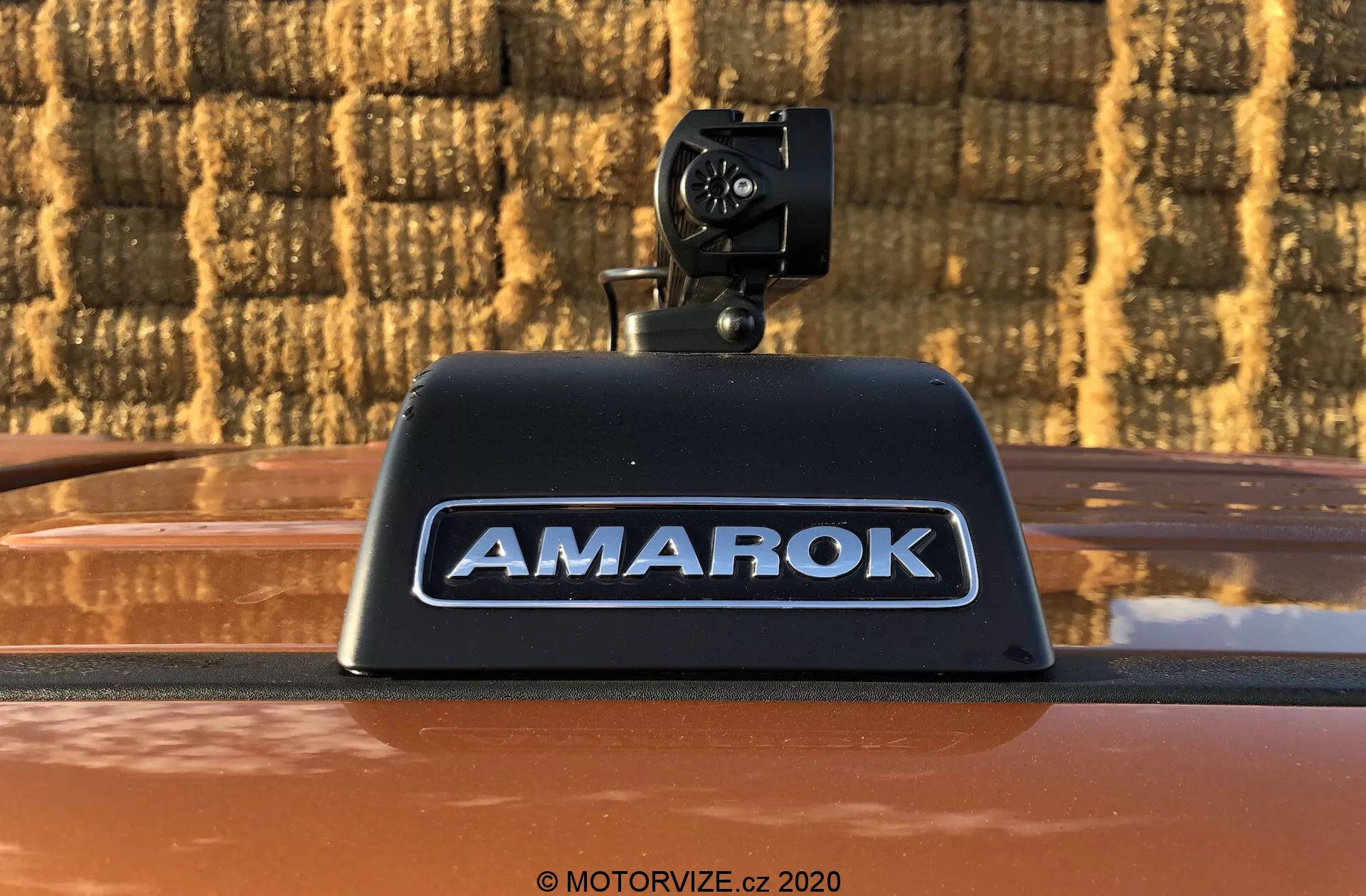 Detailná fotografia pickupu Volkswagen Amarok 2016 zobrazujúca dodatočnú spätnú kameru namontovanú na vysokomontážne brzdové svetlo s nápisom 'AMAROK' v strieborných vyrazených písmenách na kovovo bronzovej karosérii, vonku na pozadí s tehlovým vzorom.
