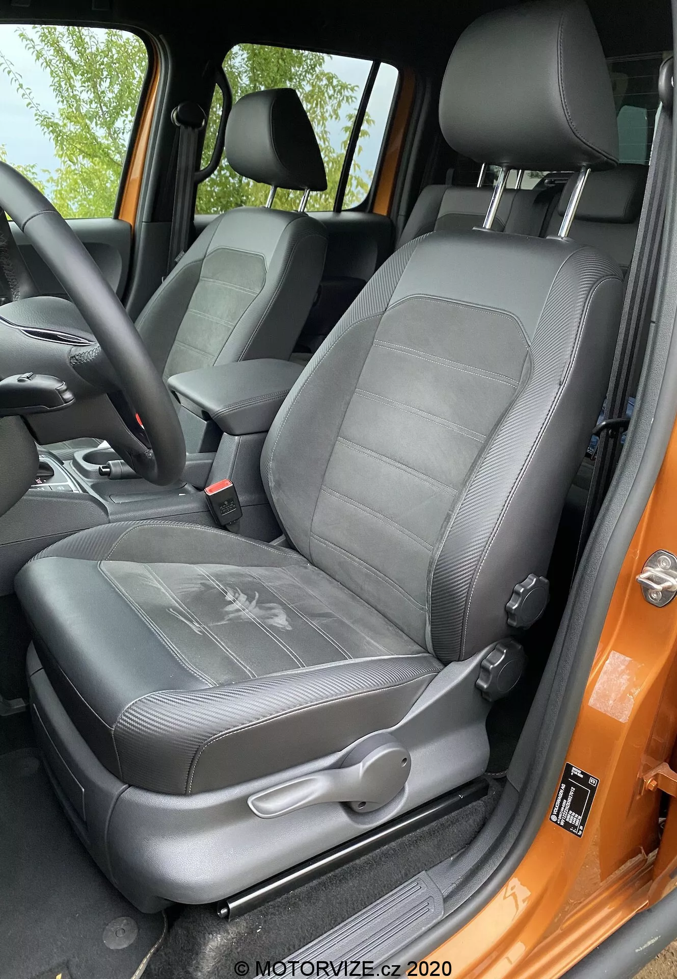 Detailný pohľad na sedadlo vodiča modelu VW-nutzfahrzeuge Amarok 2016 z pohľadu osoby stojacej vonku pri otvorených dverách. Čierne čalúnené sedadlo má vzorovaný dizajn so štruktúrovanými rebrami a integrovanú nastaviteľnú opierku hlavy. Viditeľné sú manuálne nastavovacie páčky a mechanizmus trojbodového bezpečnostného pásu. Bočná opierka naznačuje bočnú podporu a rám dverí natretý výraznou oranžovou farbou kontrastuje s čiernym interiérom. Sedadlo vyzerá byť dobre udržiavané a je osvetlené prirodzeným denným svetlom.