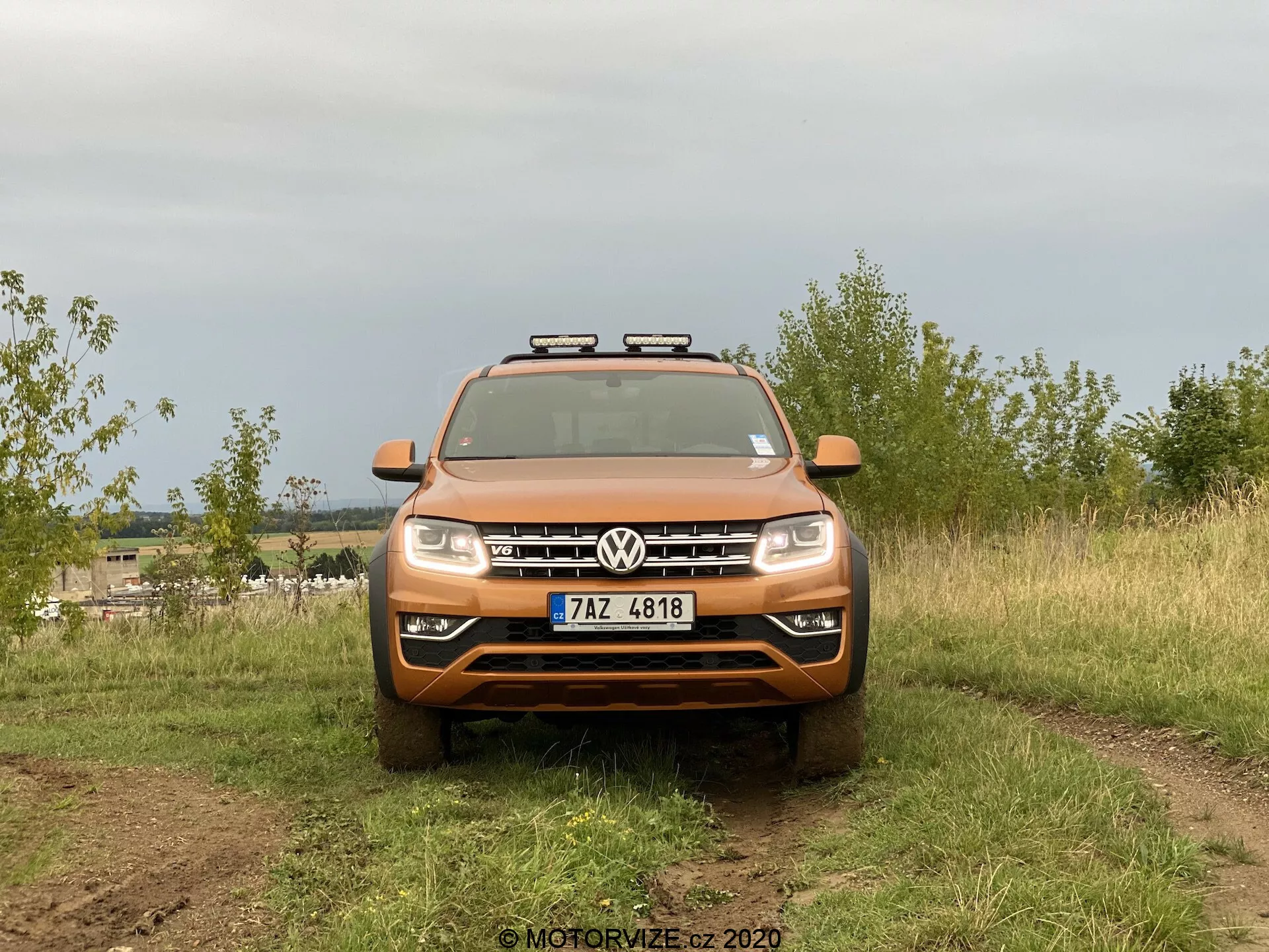 Predný pohľad na SUV Volkswagen Amarok zachytený vo vonkajšom prostredí. Oranžové vozidlo má výrazný emblém Volkswagen na mriežke, horizontálne lamely, štandardné svetlomety a prídavné svetlá na streche. Obrázok zdôrazňuje terénne schopnosti, s čiastočne viditeľnými ťažkými pneumatikami a špinou na povrchu vozidla. Je umiestnené na prírodnom teréne s rozmazaným pozadím, osvetlené rozptýleným svetlom z oblačnej oblohy.