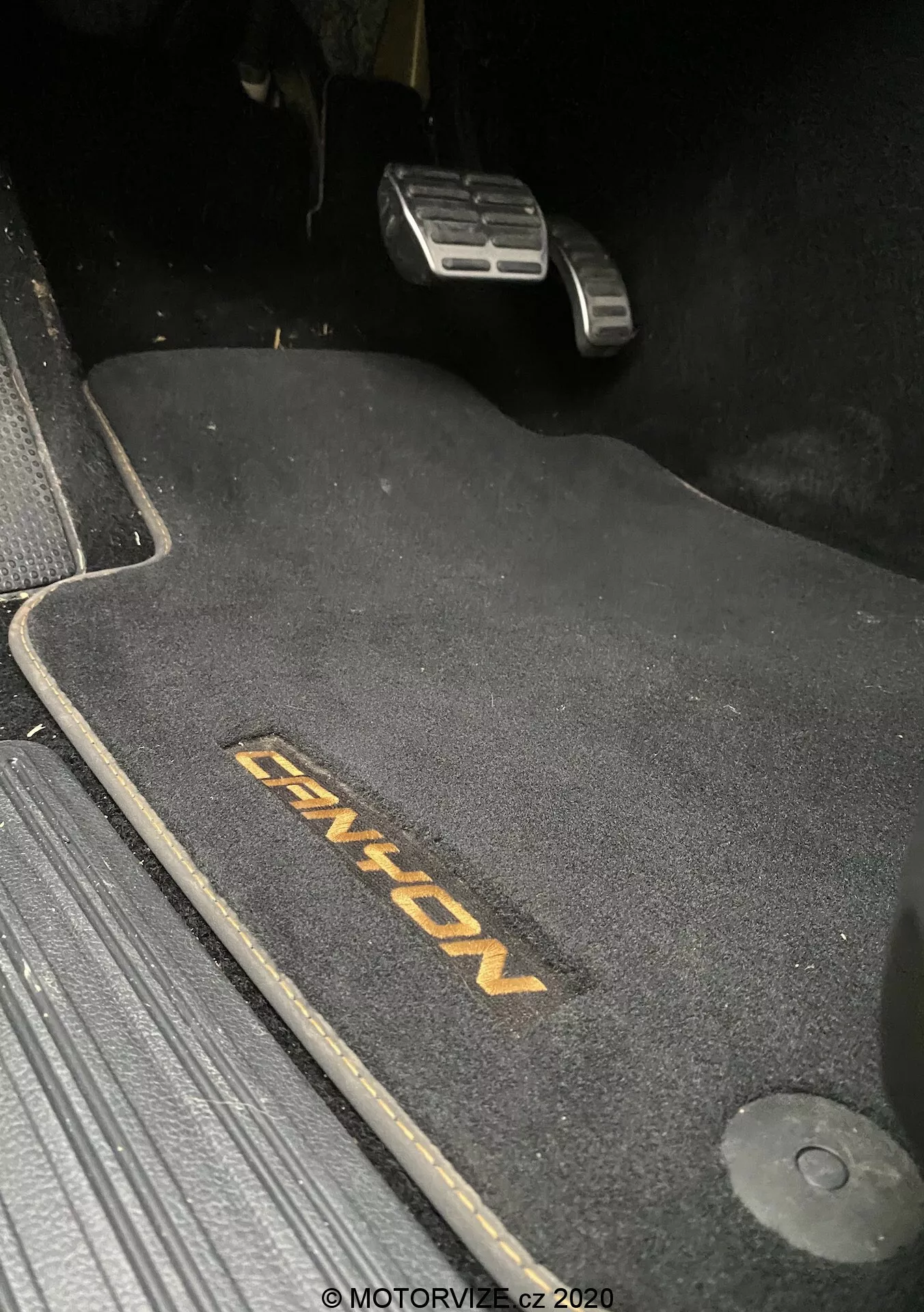 Detailný záber na oblasť podlahy na strane vodiča vo VW-Nutzfahrzeuge Amarok 2016, zobrazujúci tmavú podložku s výšivkou 'ENYON'. Viditeľné sú kovové pedále plynu a brzdy, pričom brzdový pedál je umiestnený vyššie, čo naznačuje, že je zabrzdený. Podložka je tvarovaná, aby sa prispôsobila priestoru, a na pravej strane je háčik na uchytenie, ktorý zabezpečuje, aby sa podložka neposunula. Podložka kontrastuje s tmavým okolím, čo naznačuje temnú tématiku interiéru. Scéna je slabo osvetlená, pravdepodobne vyfotografovaná v noci alebo za nízkych svetelných podmienok.