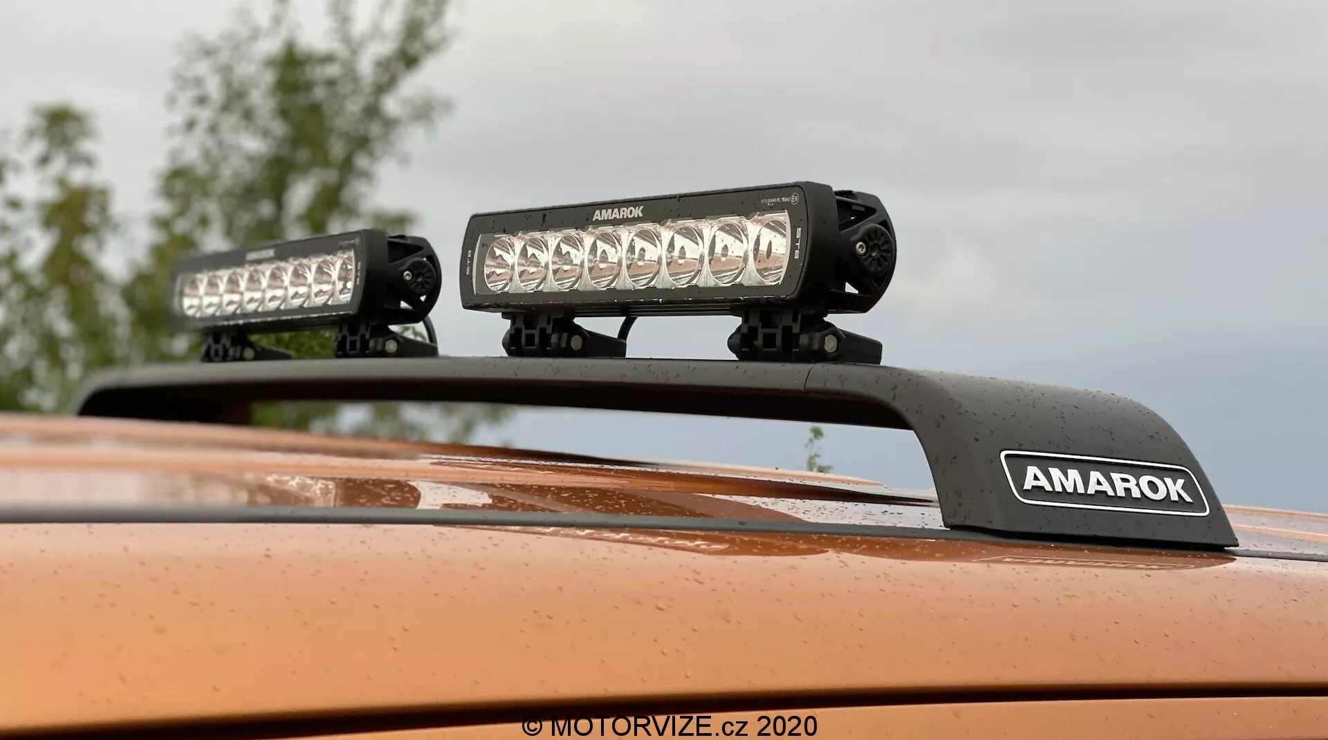 Detailný záber zo spodnej strany zameraný na strechu Volkswagenu Amarok 2016 s aftermarketovým strešným osvetlením, dvomi LED svetelnými lištami na držiaku s nápisom 'AMAROK', metalická oranžová farba; ideálne pre doplnky pre terénne vozidlá alebo osvetlenie vozidiel.