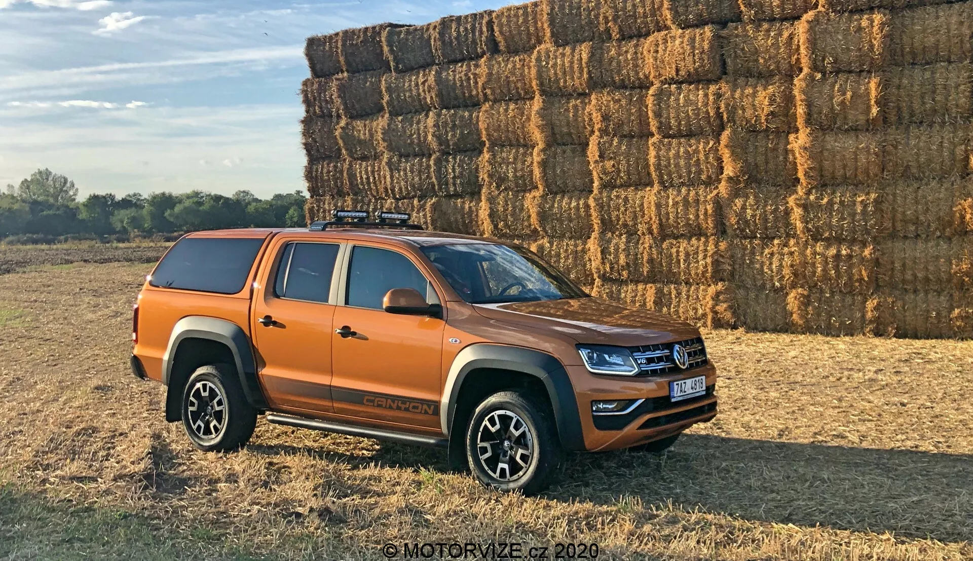Bočný pohľad na oranžový pickup Volkswagen Amarok, zaparkovaný vonku na nerovnom, potenciálne štrkovitom alebo trávnatom povrchu. Vozidlo je umiestnené pod 30-stupňovým uhlom vo vzťahu ku kamere, ukazujúc bočný profil a čiastočný pohľad na prednú mriežku a svetlomety. Moderný stredne veľký pickup má čerstvé dizajnové línie a LED osvetlenie, čo naznačuje, že ide o nedávny model. Viditeľné sú štyri dvere, čo signalizuje dvojkabinovú konfiguráciu, viacramenné zliatinové kolesá, terénne pneumatiky, tvrdý kryt na ložnom priestore vozidla a strešný nosič. Bočné zrkadlá pravdepodobne obsahujú smerovky. Fotografované v teplom dennom svetle s dlhými tieňmi, v pozadí je veľká kopa balíkov sena, čo naznačuje vidiecke prostredie.
