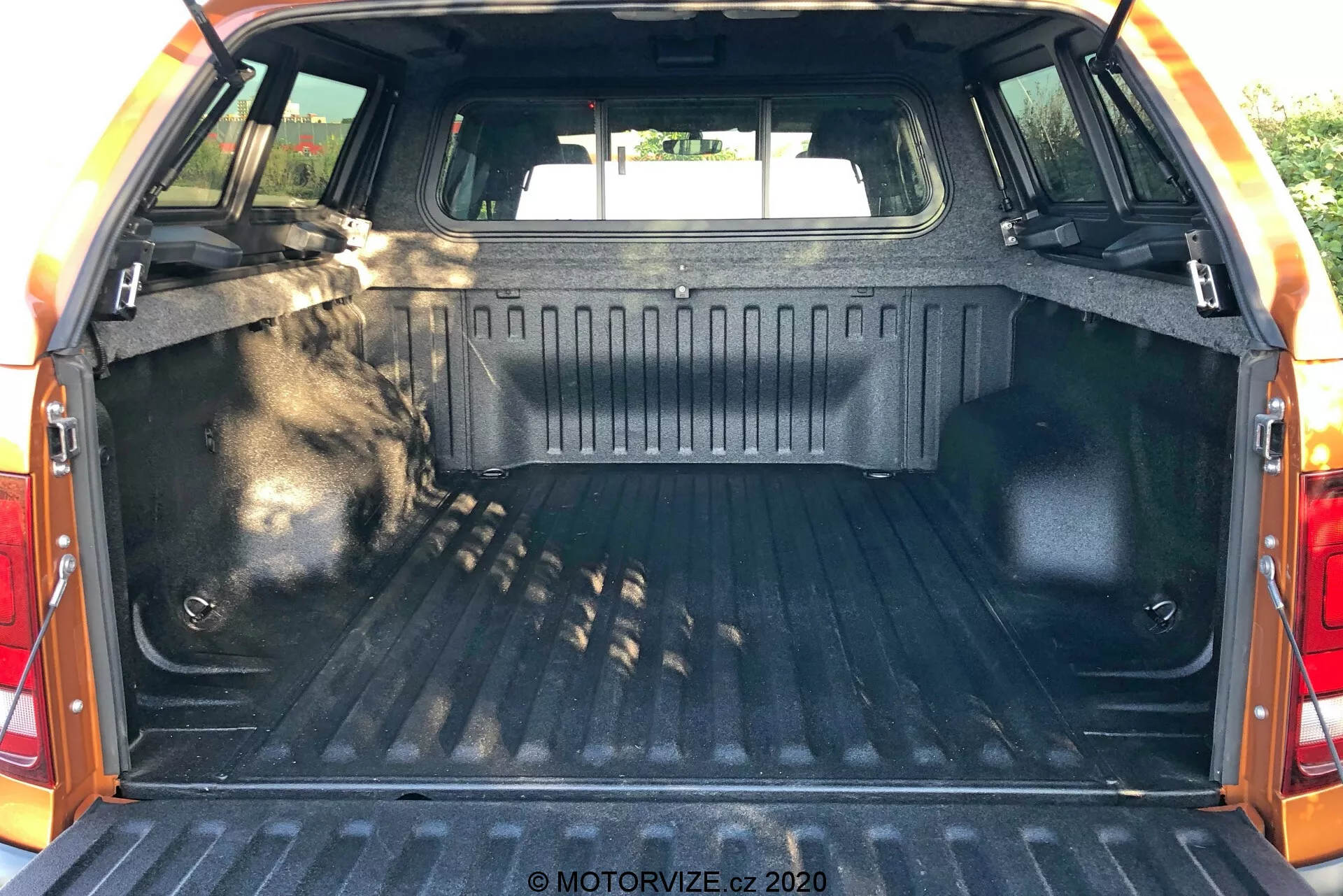 Interiér nákladného priestoru VW Nutzfahrzeuge Amarok 2016, pohľad zozadu s otvorenými zadnými dverami, ukazujúci oblasť kufra. Nákladný priestor obsahuje odolnú vzorovanú výstelku, bočné steny dokončené odolným materiálom, upevňovacie body na zabezpečenie nákladu, vnútorné osvetlenie pre viditeľnosť, zadné sedadlá viditeľné v prednej časti priestoru a ochranný lem na okraji zadných dverí.