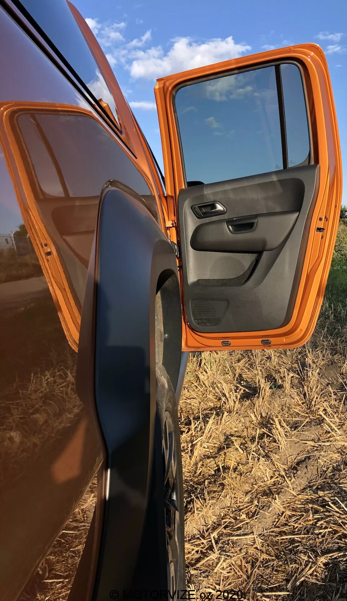 Detailný pohľad na čiastočne otvorené predné dvere spolujazdca oranžového VW Nutzfahrzeuge Amarok 2016, zvýrazňujúci jasne oranžovú farbu exteriéru a kontrastný čierny vnútorný panel dverí s integrovanými ovládacími prvkami. Auto je zaparkované diagonálne, čím predstavuje bočný profil s dverami otváranými v uhle 70 stupňov, zdôrazňujúc zrkadlo s čiernym krytom. Odrazy na dverách ukazujú jasnú oblohu a rovinatú poľnohospodársku krajinu, zdôrazňujúc vidiecke prostredie.