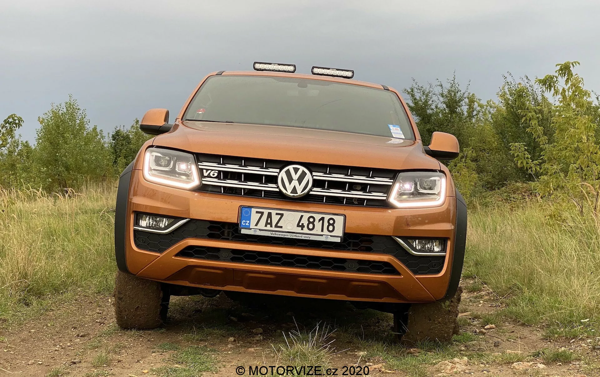 Predný pohľad na kovovo oranžový pickup Volkswagen Amarok, ktorý ukazuje svoju robustnú postavu a terénne schopnosti s výraznou mriežkou s emblémom VW a odznakom V6. Obrázok, nasnímaný z nízkeho uhla, zachytáva celú šírku prednej časti vozidla, vrátane svetlometov a prídavných svetiel pre terénne jazdenie, s prírodným pozadím poľa a stromov, ktoré zvýrazňujú robustnosť pickupu. Viditeľné prvky zahŕňajú pevný predný nárazník, kryciu dosku, aktívny pokročilý osvetľovací systém a evidenčnú značku Českej republiky.