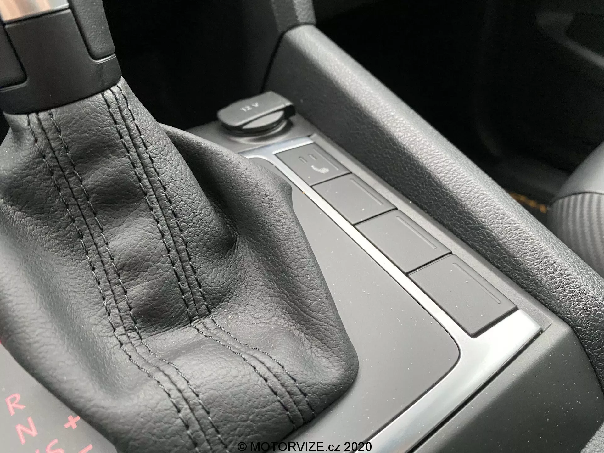 Detailný záber na stredovú konzolu vozidla VW Nutzfahrzeuge Amarok 2016, zameriavajúci sa na oblasť radiacej páky. Hlavným prvkom je radiaca páka, potiahnutá čiernou kožou s viditeľným šitím. Písmená 'PRNDS' za radiacou pákou označujú automatickú prevodovku. Napravo sú štyri štvorcové tlačidlá zarovnané s povrchom konzoly, pravdepodobne ovládajúce rôzne funkcie vozidla. Oblasť za tlačidlami môže byť malý úložný priestor. Materiály a kvalita vyhotovenia naznačujú vysokú úroveň remeselného spracovania. Ambientné svetlo z ľavého horného rohu zvýrazňuje detaily.