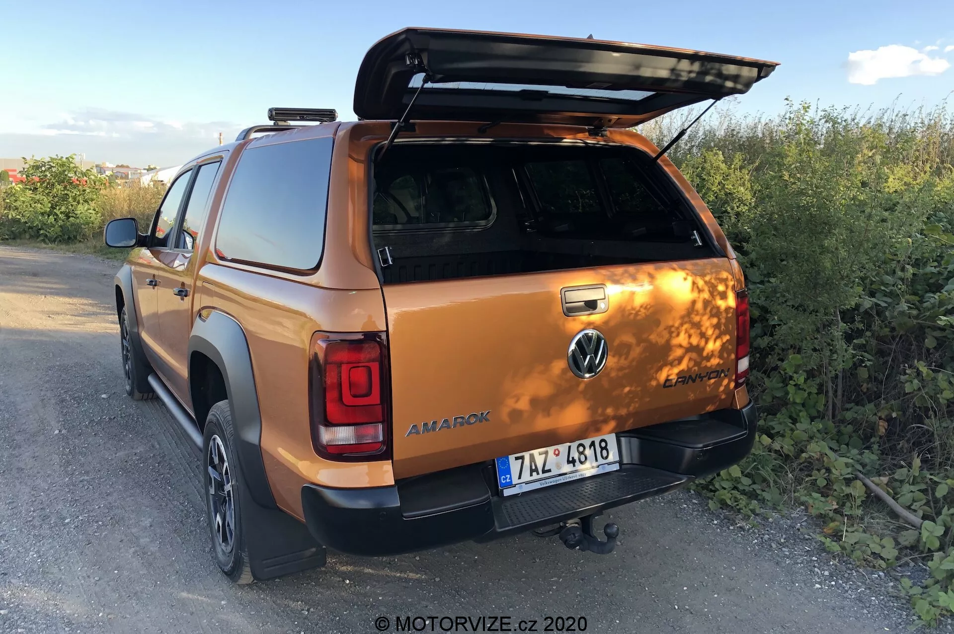 Zadný trojštvrťový pohľad na Volkswagen Amarok 2016 v metalickej oranžovej, zdôrazňujúci otvorený kryt postele, ťažné zariadenie, dvere druhej rady dvojitej kabíny a časť pravého zadného kolesa a nárazníka, zasadený do vidieckeho vonkajšieho prostredia na súmraku. Foto od MOTORVIZE.cz, 2020.