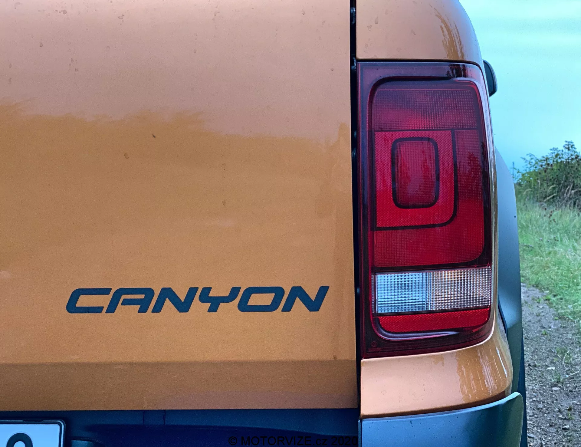 Detailný pohľad na pravý zadný roh vozidla VW Nutzfahrzeuge Amarok 2016. Na zadných dverách je viditeľný znak 'CANYON' medzi známkami ľahkého znečistenia, čo naznačuje použitie na nespevnených cestách. Fotografia zvýrazňuje predovšetkým moderný obdĺžnikový svetelný klaster so sekciami pre brzdové, spätné a pravdepodobne smerové svetlá. Snímka urobená z približne 45-stupňového bočného uhla zachytáva čiastočne viditeľnú karosériu vozidla s vidieckym alebo polo-vidieckym pozadím.