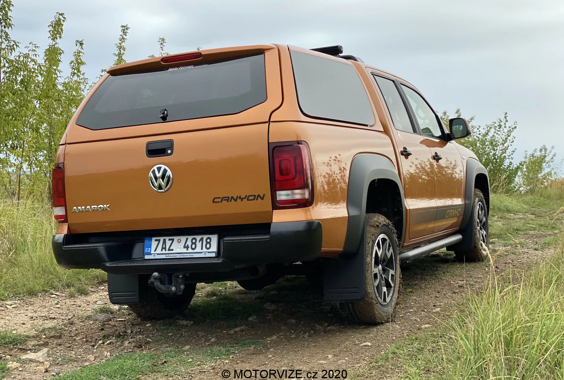 Profesionálny a podrobný pohľad na tri štvrtiny zadnej časti Volkswagen Amarok 2016, zaparkovaného na mierne nerovnom teréne s trávou. Zvýraznená je pravá zadná strana pickupu, ktorá ukazuje uzavretý kryt ložného priestoru v oranžovej alebo medenej farbe. Zadné dvere majú logo Volkswagen a názov modelu 'AMAROK'. Zadné svetlá majú moderný segmentovaný dizajn a zadný nárazník obsahuje stredný krok, pravdepodobne vybavený parkovacími senzormi. ŠPZ má chrómový alebo leštený rám a pod nárazníkom je viditeľné ťažné zariadenie, čo naznačuje schopnosť ťahať. Terénne pneumatiky a viacramenné hliníkové kolesá naznačujú prípravu na off-road a vyššiu úroveň vybavenia. Na spodnej strane ložného priestoru sú viditeľné čierne ochranné opláštenia.