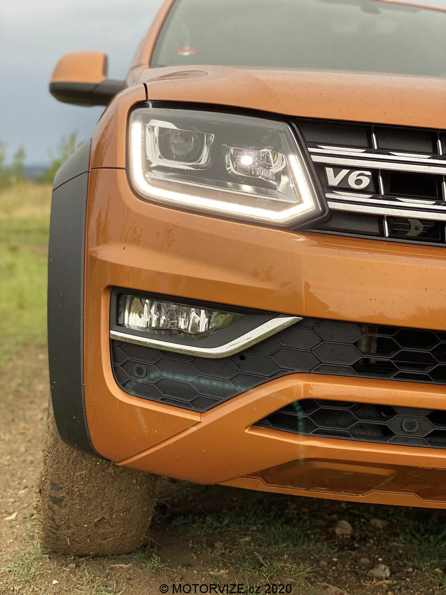 Detailný záber na ľavý predný roh oranžového VW Nutzfahrzeuge Amarok 2016, zobrazujúci svetlomet s moderným čírym krytom a LED technológiou, hmlové svetlá v čiernej mriežke, čiastočne viditeľnú mriežku s odznakom V6, nárazník s čiernou voštinovou štruktúrou, tvarovanú kapotu a čiastočne viditeľné veľké koleso, s rozmazaným pozadím zdôrazňujúcim textúru a robustný dizajn vozidla