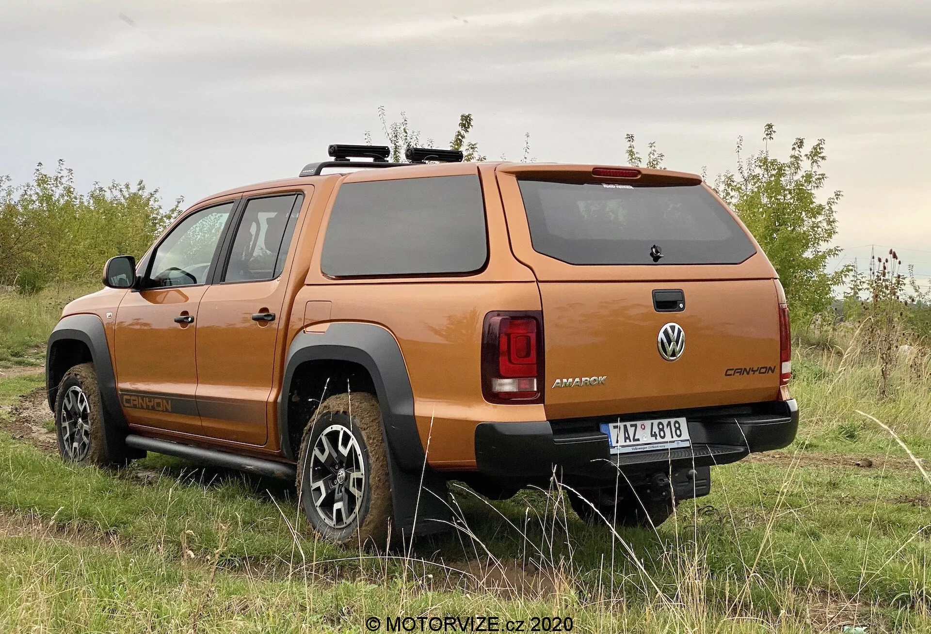 Trojštvrťový pohľad zozadu na oranžové pickup vozidlo Volkswagen Amarok 2016, ktoré zobrazuje dizajn kolies, bočný profil, zadné okno, zadné svetlá, robustný zadný nárazník a pevný kryt nákladového priestoru, zaparkované na neupravenom povrchu s vegetáciou, zdôrazňujúc jeho terénne schopnosti.