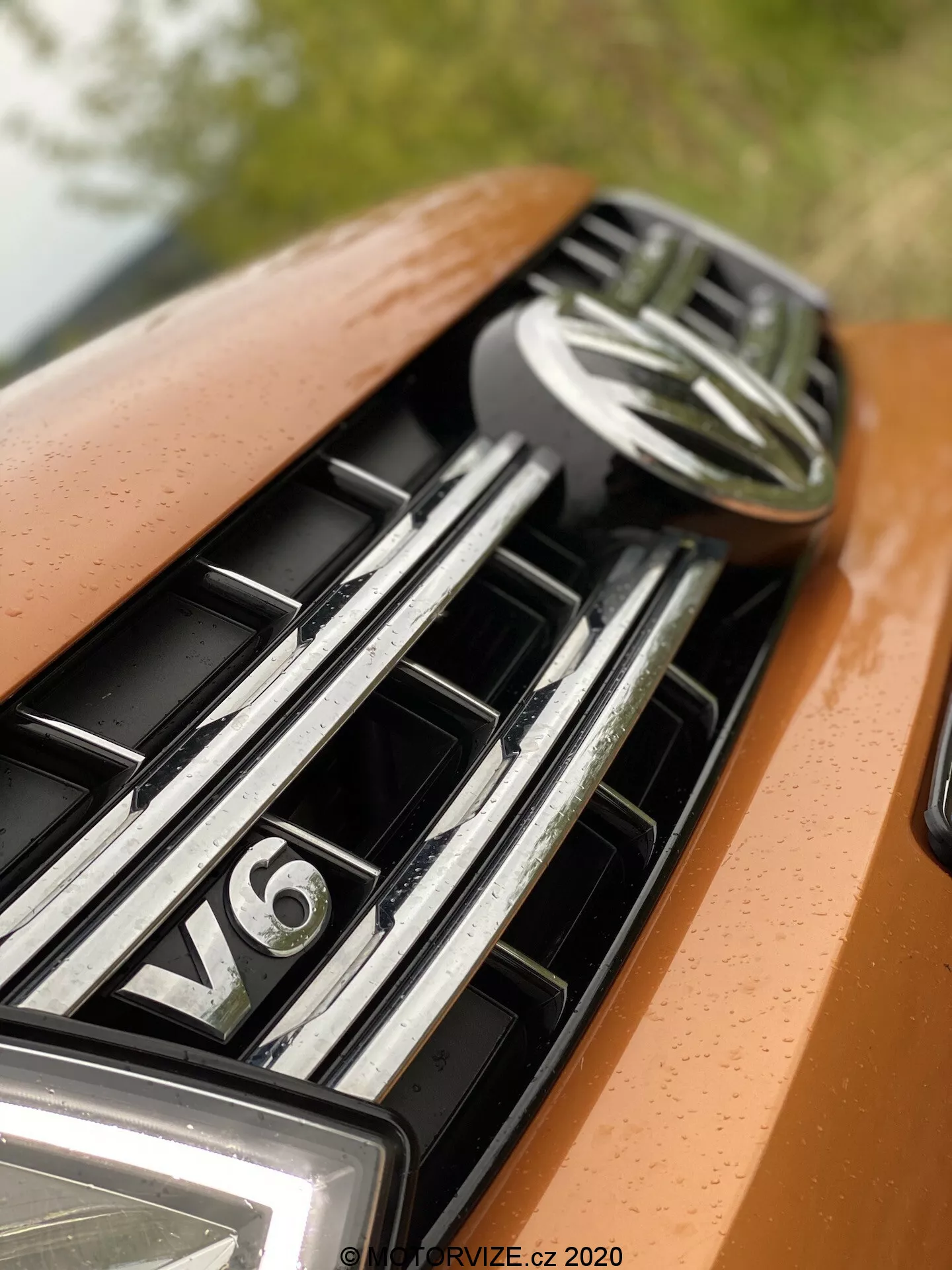 Detailný záber na kapotu a mriežku auta, zameraný na hornú časť s centrálnym emblémom a znak motora V6. Mriežka obsahuje vodorovné, lesklé chrómové lamely, čo naznačuje luxusné vozidlo. Lak auta má kovovú oranžovú alebo bronzovú farbu a mäkké, rozptýlené osvetlenie zvýrazňujúce hladký dizajn a prémiový vzhľad.