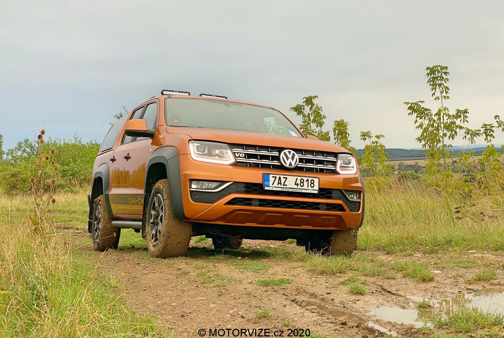 Nízky predný trojštvrťový uhol Volkswagenu Amarok 2016 na bahnitom poli, ktorý zobrazuje jeho oranžový náter, prednú mriežku s odznakom Volkswagen, svetlomety, dizajn kolies a schopnosti v teréne. Robustnosť vozidla je zvýraznená blatom na pneumatikách a spodných stranách, predné kolesá sú natočené doľava, čo zdôrazňuje vzory behúňa a výšku jazdy. Na mäkko osvetlenom pozadí, buď ráno alebo neskoro popoludní, obrázok zvýrazňuje využiteľnosť Amaroku v teréne a jeho pokročilý osvetľovací systém. Evidenčné číslo je viditeľné a pre súkromie by malo byť zakryté.