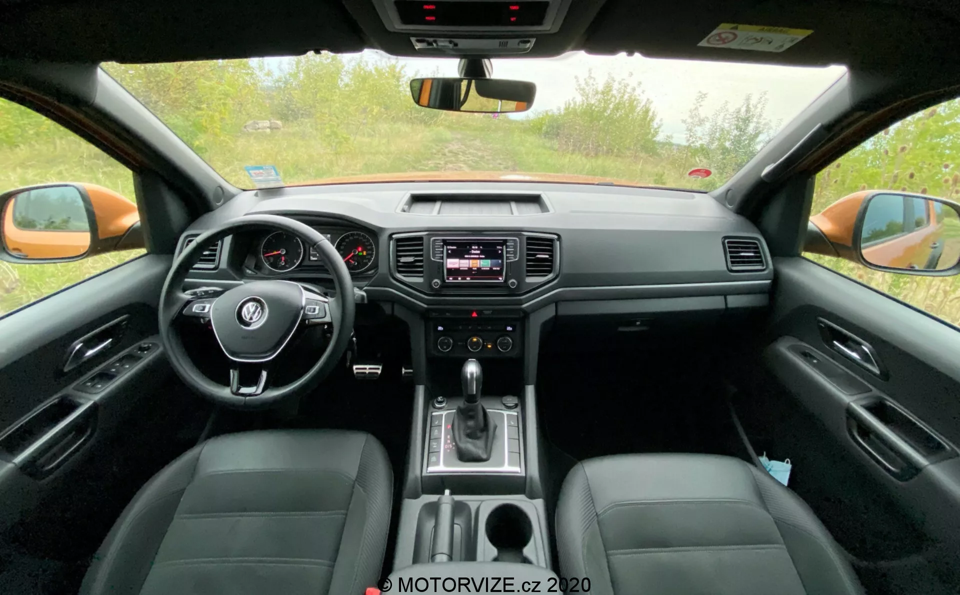 Interiérový pohľad na kabínu vozidla VW-Nutzfahrzeuge Amarok 2016. Fotografované z pohľadu zadného sedadla, tento obrázok zobrazuje stredový volant s integrovanými ovládacími prvkami, analógový prístrojový panel s malým digitálnym displejom, infotainmentový systém v strede prístrojovej dosky s ovládaním audia a klimatizácie, radiacu páku indikujúcu automatickú prevodovku a kožené sedadlá. Obrázok tiež zachytáva spätné zrkadlo, vonkajšie spätné zrkadlá a predné bočné okná, čím zdôrazňuje technologické a komfortné prvky.