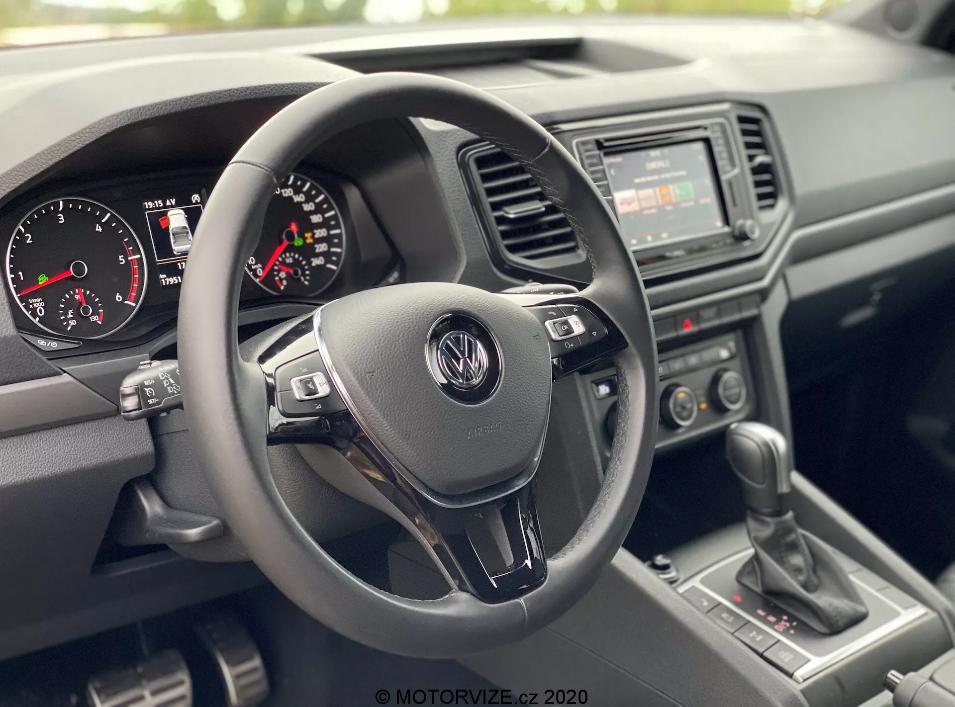 Pohľad do interiéru VW Nutzfahrzeuge Amarok 2016 z perspektívy predného spolujazdca, zobrazujúci oblasť kokpitu vodiča. Ústredný bod sa zameriava na trojramenný volant s logom Volkswagen a multifunkčnými ovládacími tlačidlami, prístrojový panel s analógovým rýchlomerom, otáčkomerom a malým digitálnym displejom, základné rádio s manuálnymi prepínačmi klimatizácie na stredovej konzole, manuálnu radiacu páku a textúrované plastové materiály pre prístrojovú dosku a dvere, zdôrazňujúci odolný a praktický dizajn.