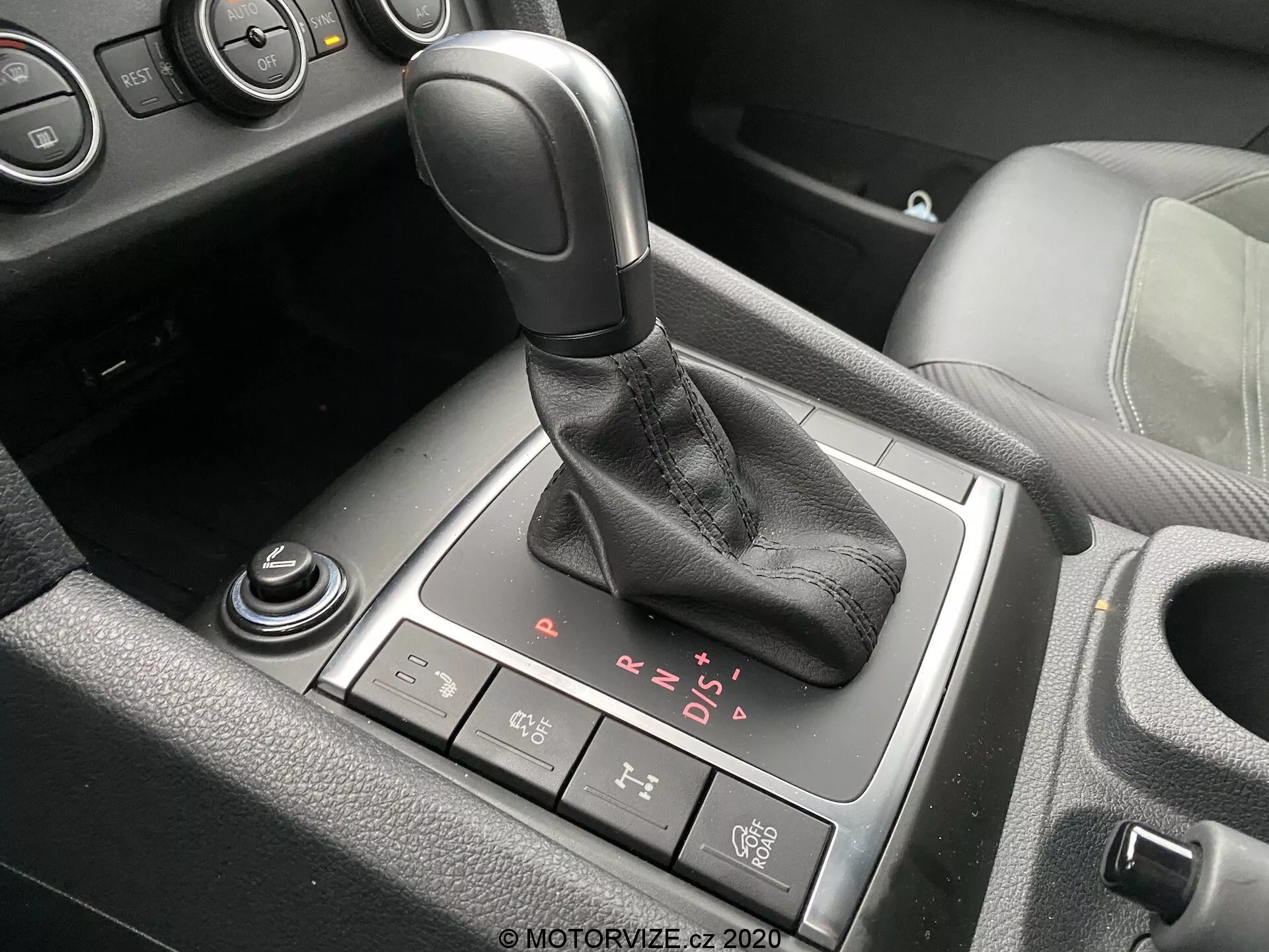 Detailný pohľad na oblasť radiacej páky v interiéri VW Nutzfahrzeuge Amarok 2016. Automatická radiaca páka s čiernou rukoväťou a koženým návlekom je ústredne umiestnená na obrázku s viditeľnými možnosťami P, R, N, D a sekvenčným manuálnym režimom. Tlačidlo elektronickej parkovacej brzdy sa nachádza za radiacou pákou, zatiaľ čo čiastočne viditeľný otočný volič a ďalší volič sa nachádzajú vpredu a v ľavom dolnom rohu. Sú viditeľné aj čiastočné pohľady na držiak pohárov a stredovú konzolu s čiernymi plastovými a pravdepodobne hliníkovými alebo chrómovými akcentmi, čo naznačuje moderné vozidlo so strednou alebo vyššou úrovňou interiéru.