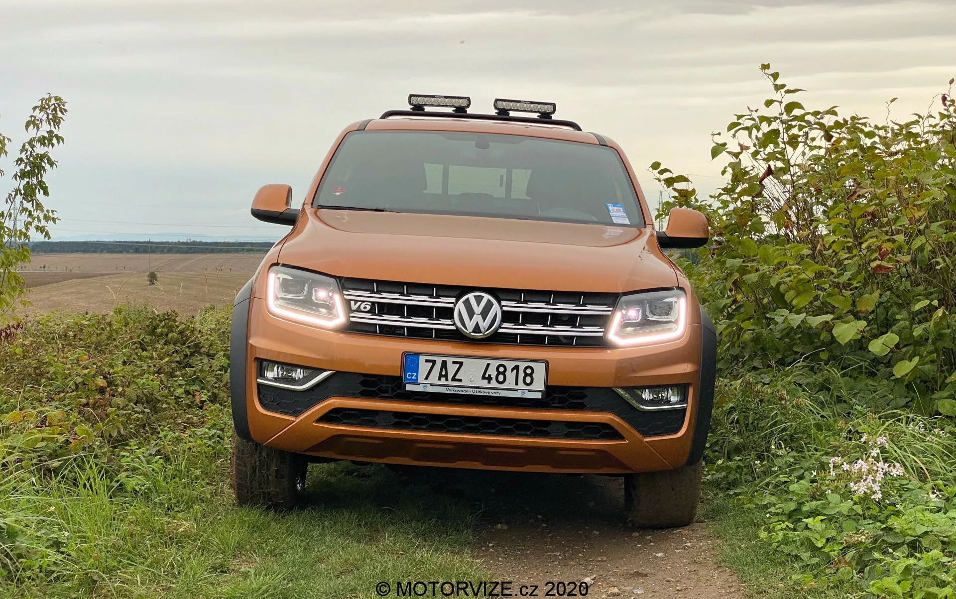 Pohľad spredu na Volkswagen Amarok SUV v metalickej oranžovej farbe, zaparkovaný na tráve s vidieckym off-road pozadím, ukazujúc symetrickú masku, svetlomety, chrómové akcenty a pomocné strešné svetlá.