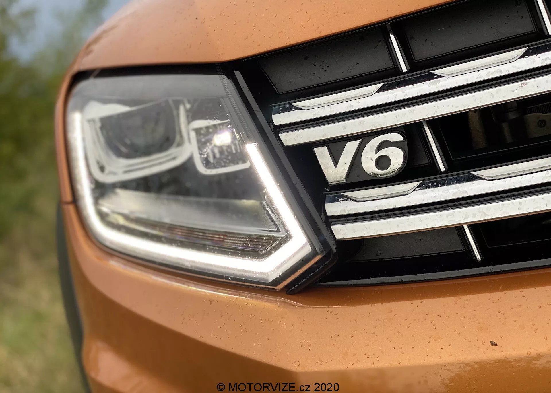 Detailný záber na prednú ľavú časť vozidla VW Nutzfahrzeuge Amarok 2016. Obrázok zachytáva moderný, hranatý svetlomet s LED denným svietením a technológiou projektorových svetiel, spolu s chrómovými detailmi. Maska chladiča má horizontálne chrómové pásy a odznak 'V6', indikujúci šesťvalcový motor vozidla. Časť kapoty je vo výraznej oranžovej farbe, čo naznačuje športový dizajn.