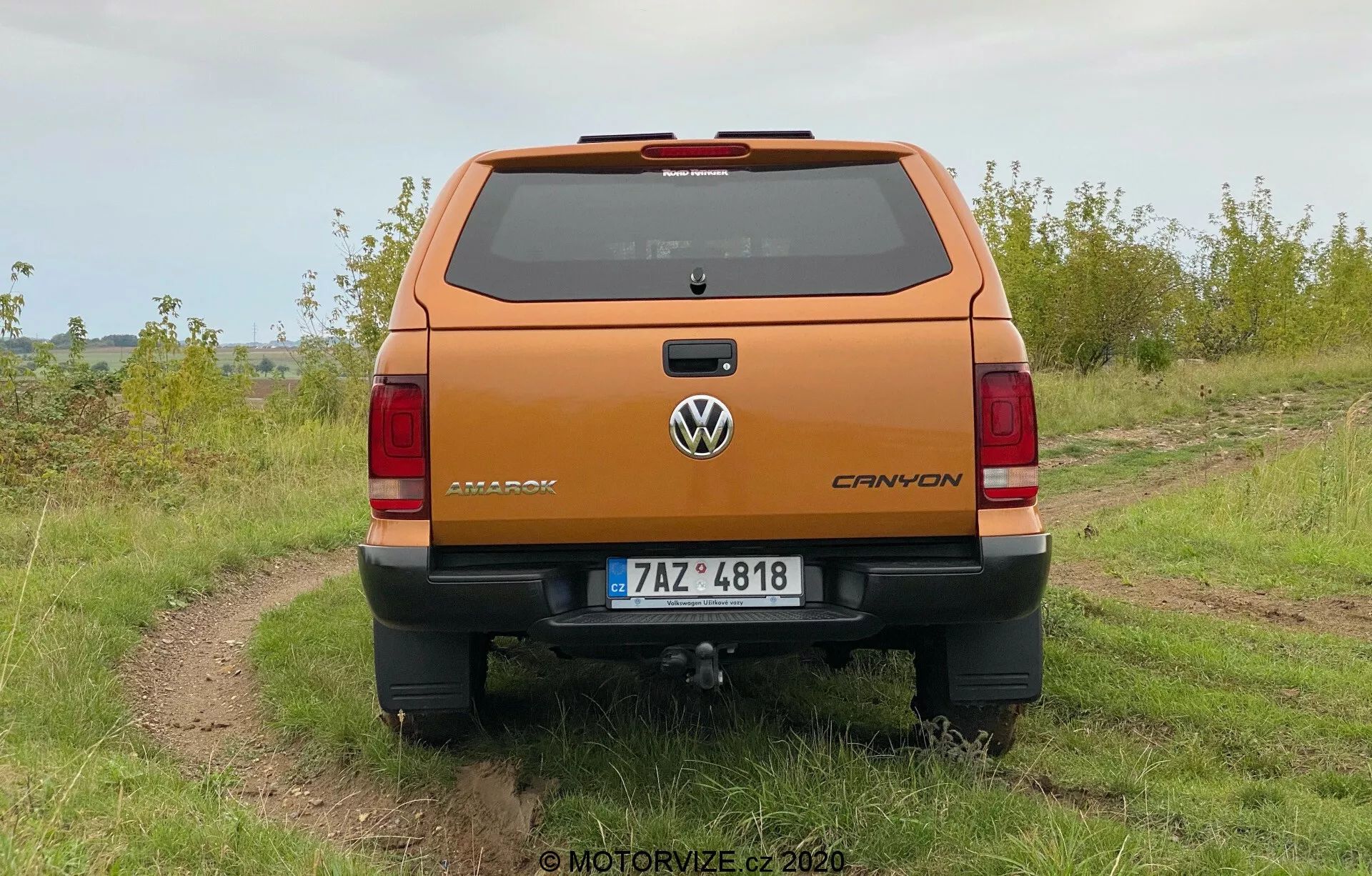 Zadný pohľad na oranžový metalický Volkswagen Amarok 2016 pickup zaparkovaný vonku na nerovnom štrkovom chodníku s trávou po oboch stranách pod zamračenou oblohou. Fotografia zobrazuje jasnú perspektívu zadnej strany vozidla, vrátane znaku Volkswagen, názvu Amarok a emblému V6. Vozidlo má zadný nárazník s integrovaným schodom, centrálne umiestnenú tabuľku s evidenčným číslom a viditeľné ťažné zariadenie. Viditeľné sú vertikálne orientované zadné svetlá a tretie brzdové svetlo na strešnej línii nad zadným oknom, ako aj odmrazovacie línie.