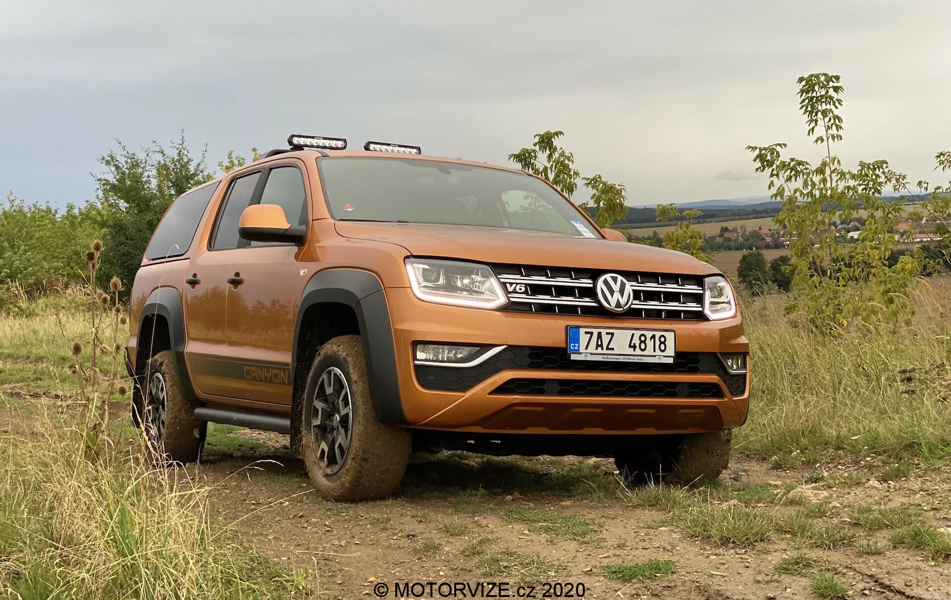 Obrázok zobrazuje moderné SUV, model Volkswagen Amarok 2016, zaparkovaný vonku na nerovnom teréne, ktorý demonštruje jeho schopnosti jazdy v teréne. Pohľad z nízkeho predného uhla zobrazuje prednú štvrtinu vozidla pokrytú bahennými zvyškami z nedávneho používania v teréne. Široká predná mriežka s charakteristickým znakom VW je lemovaná obdĺžnikovými svetlometmi s LED denným osvetlením. Pod mriežkou sa nachádza robustný predný nárazník striebornou ochrannou doskou na ochranu podvozku. Kontúrovaná kapota pridáva SUV mohutný vzhľad. Viditeľné sú strešné nosiče, ktoré naznačujú možnosti prepravy ďalšieho nákladu. Kolesá, čiastočne zakryté blatom, majú hlboký dezén pre lepšiu trakciu v náročnom teréne. Viditeľný je výrazný oblúk nad predným kolesom a bočný schodík uľahčujúci vstupl a vystupovanie z vozidla. Zreteľné sú aj strešné osvetlenie a bočné zrkadlá. Auto je umiestnené do pozadia s vegetáciou a kopcovitým terénom pod zamračenou oblohou, čo zvýrazňuje jeho terénne schopnosti a vonkajšiu príťažlivosť. Ľavý volant a viditeľná registračná značka naznačujú, že vozidlo sa nachádza v krajine s pravostranným jazdením. Tento záber zvýrazňuje dizajn auta, terénne prvky a moderné osvetľovacie technológie.