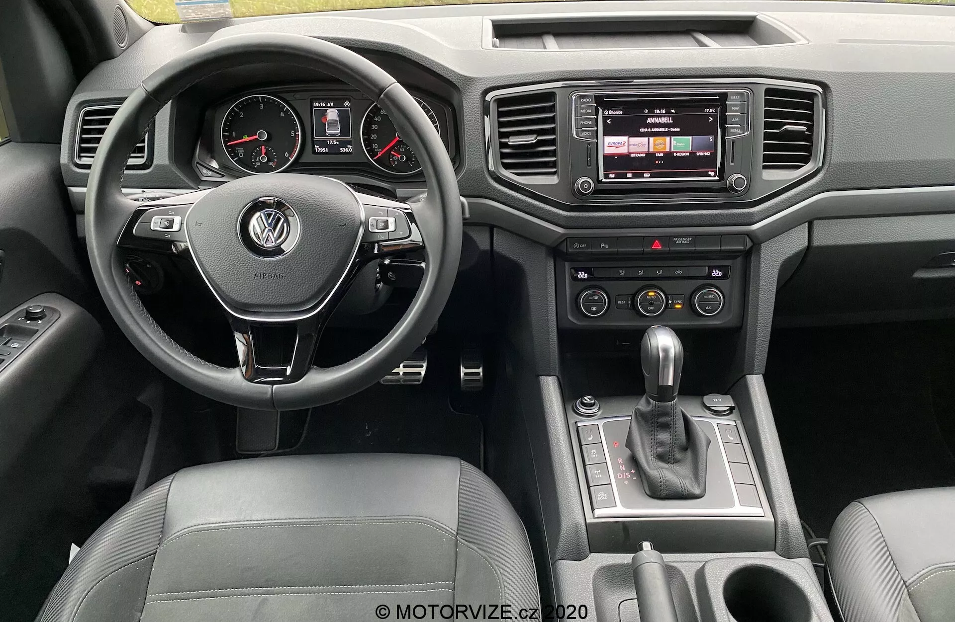 Pohľad na prístrojovú dosku Volkswagen Amarok 2016 z pohľadu vodiča. Centrálne umiestnený multifunkčný volant potiahnutý kožou s logom VW, analógovými ukazovateľmi a digitálnym displejom na prístrojovej doske. Stredová konzola obsahuje dotykový infotainment systém, tlačidlá klimatizácie a automatickú prevodovú páku. Luxusné detaily zahŕňajú kožené a chrómové povrchy, s viditeľnými ovládacími prvkami na dverách.